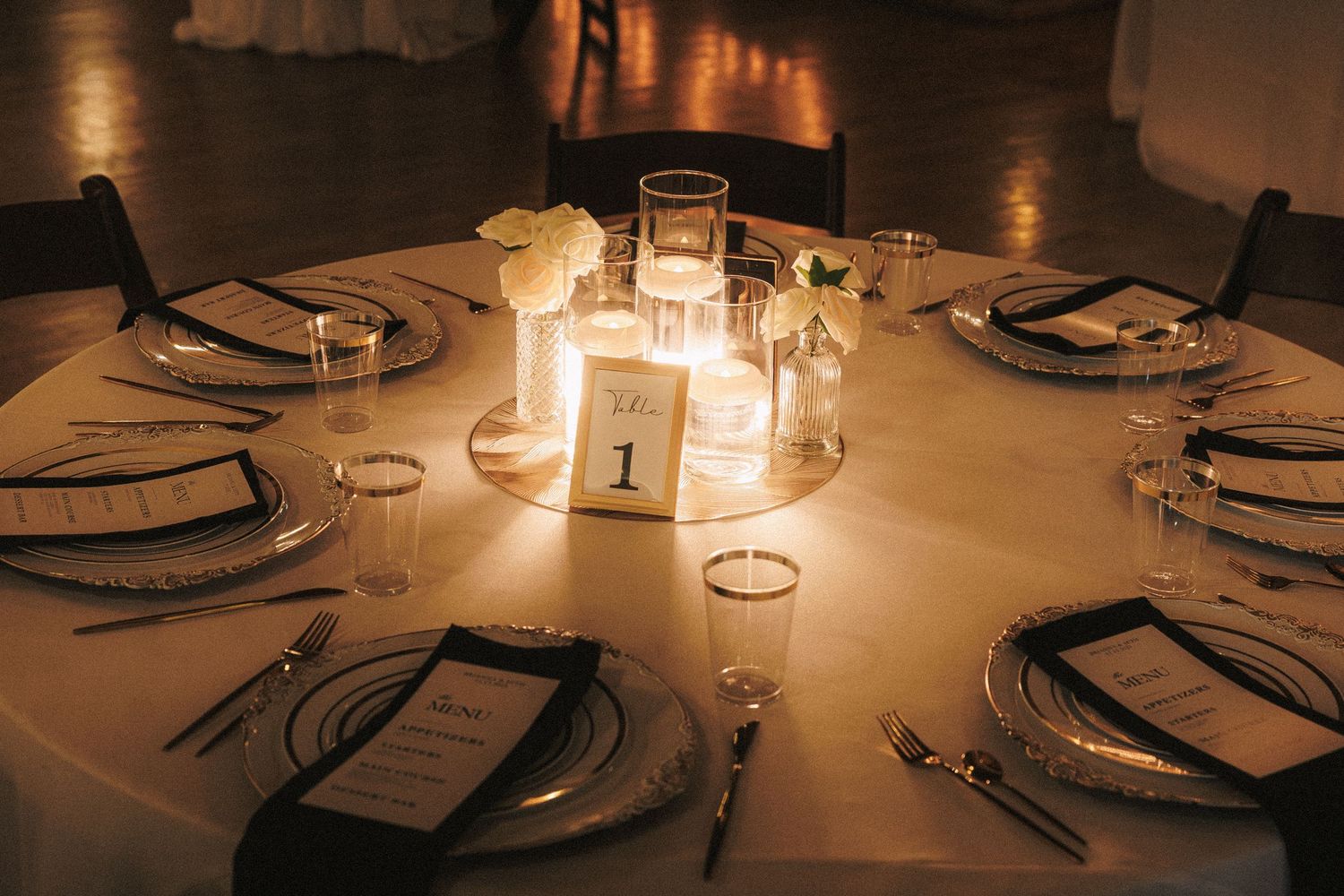 Elegant candlelit wedding reception table with white florals, crystal glasses, and black menu cards for table number one.