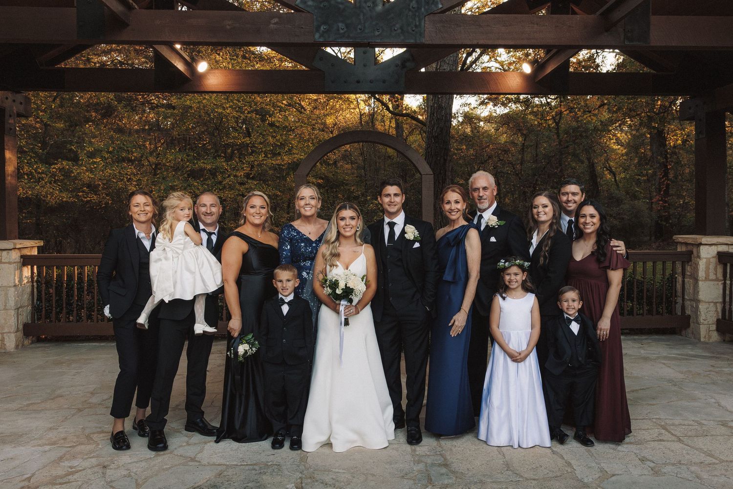 Large wedding family portrait group posed under lit pavilion surrounded by autumn foliage at dusk.