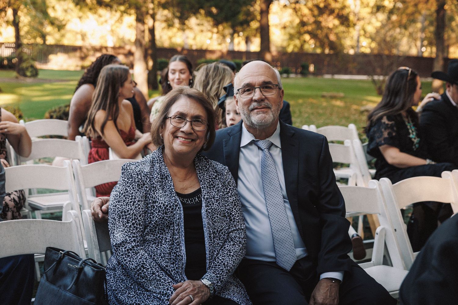 Smiling couple seated in white chairs at outdoor wedding ceremony surrounded by guests.