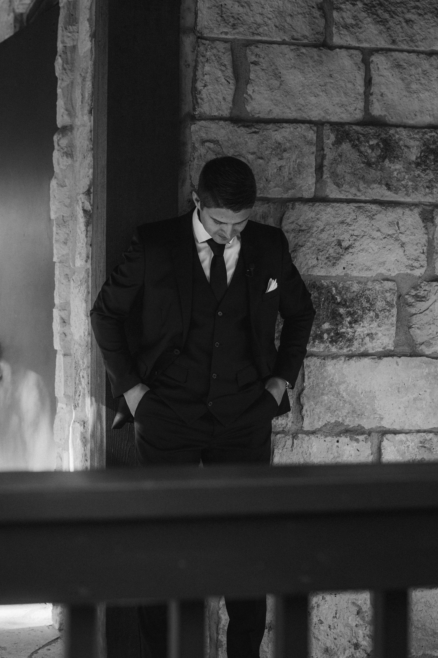 Black and white portrait of groom in three-piece suit standing pensively against stone wall exterior.
