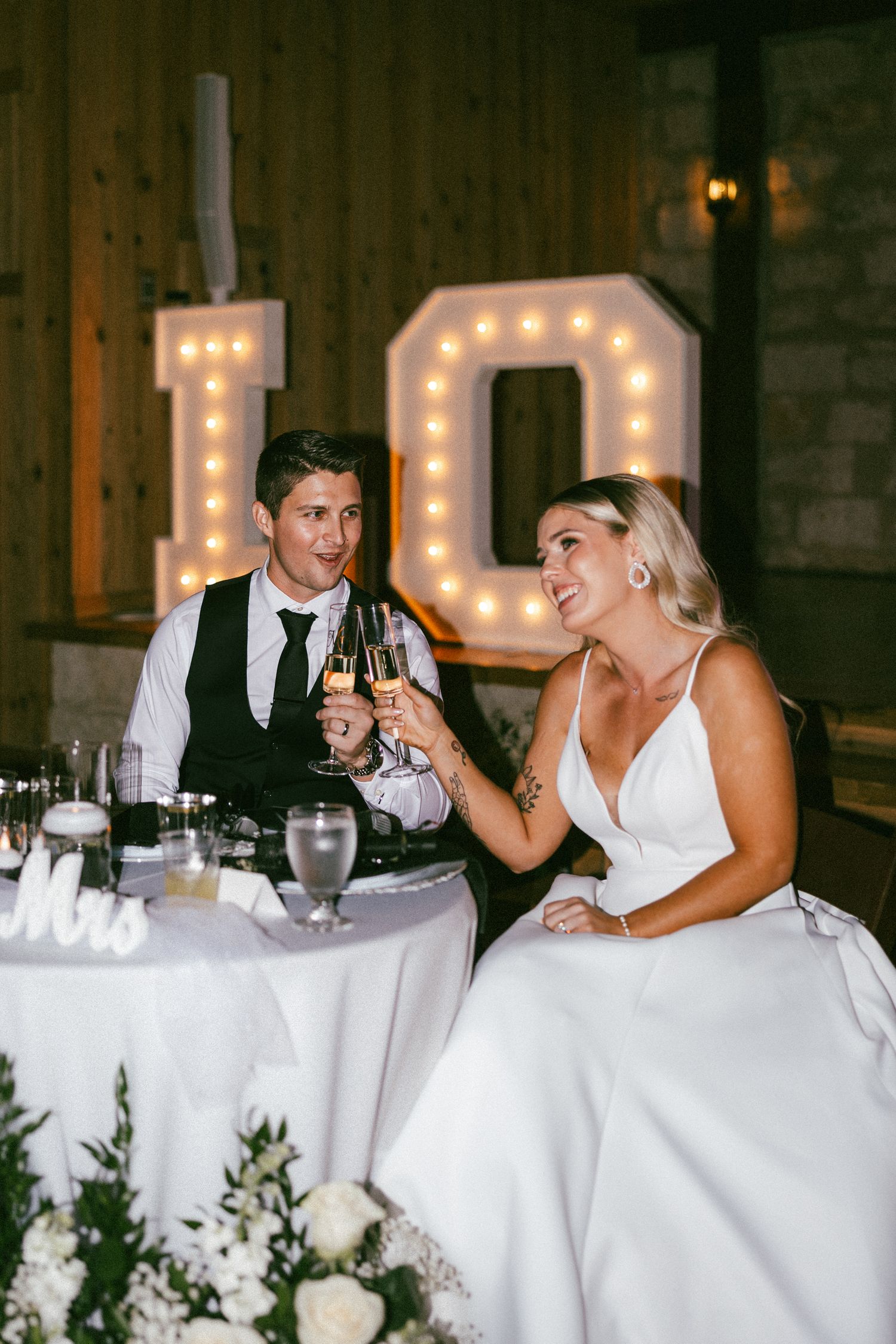 Bride and groom toasting champagne at sweetheart table with illuminated IO marquee letters.