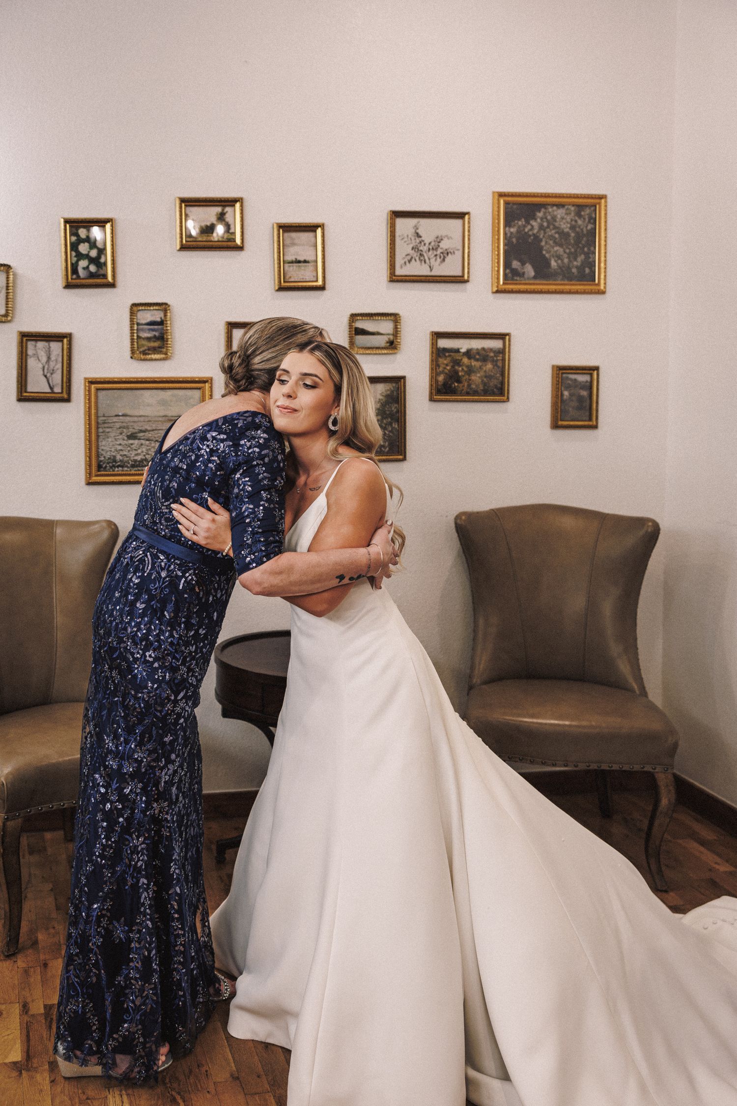 A bride in a white gown shares an emotional hug with a woman in a navy sequin dress before the wedding.