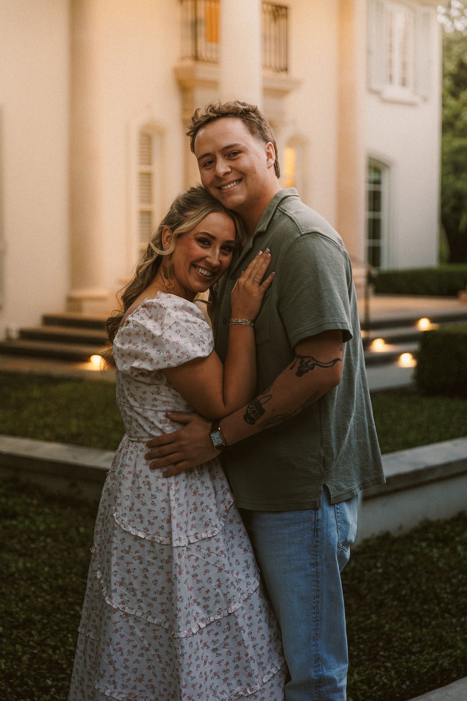 Smiling couple embracing in front of an elegant white mansion at golden hour.