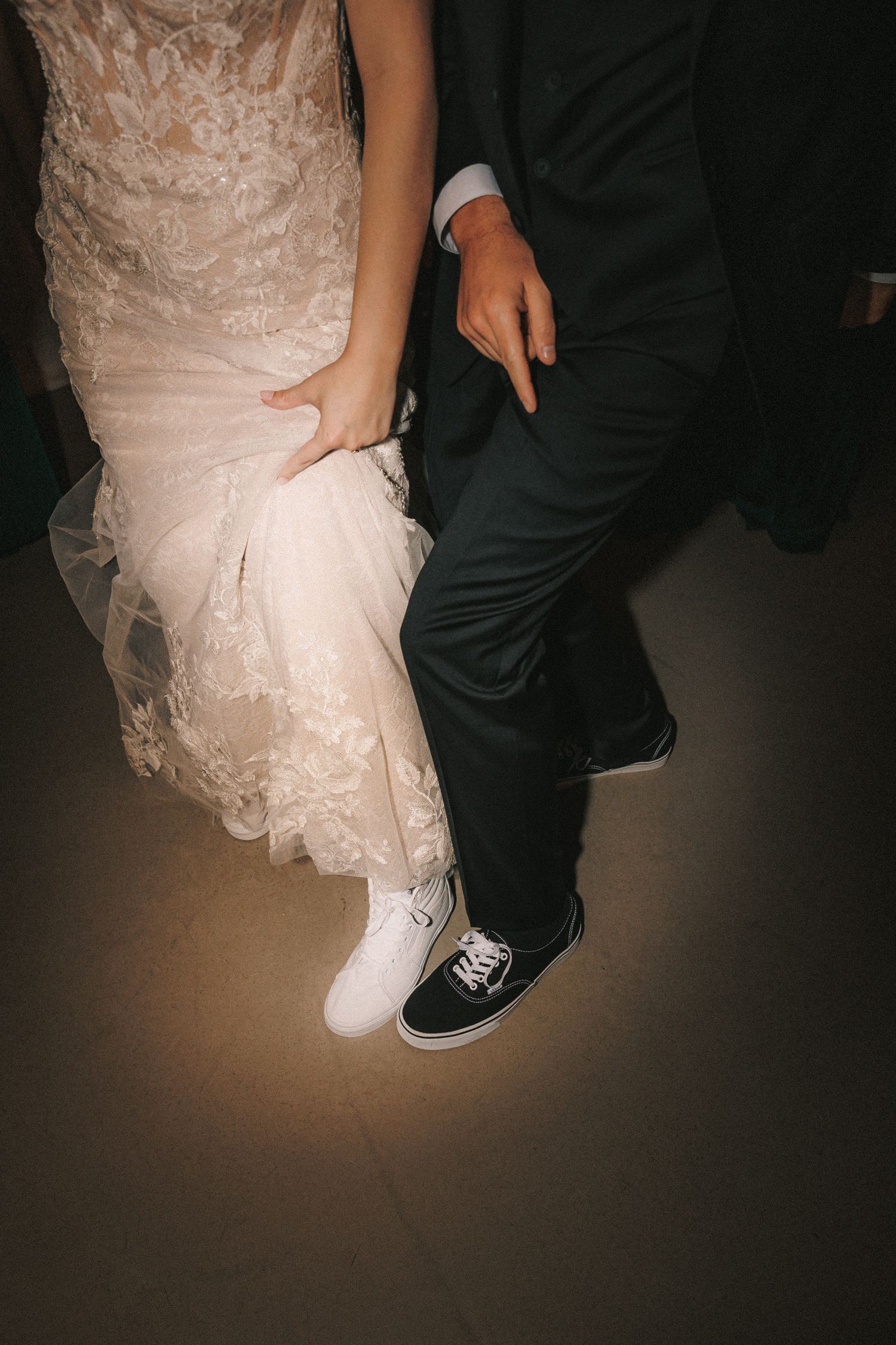 Close-up of bride in lace gown and groom in suit wearing black and white Vans sneakers while dancing.