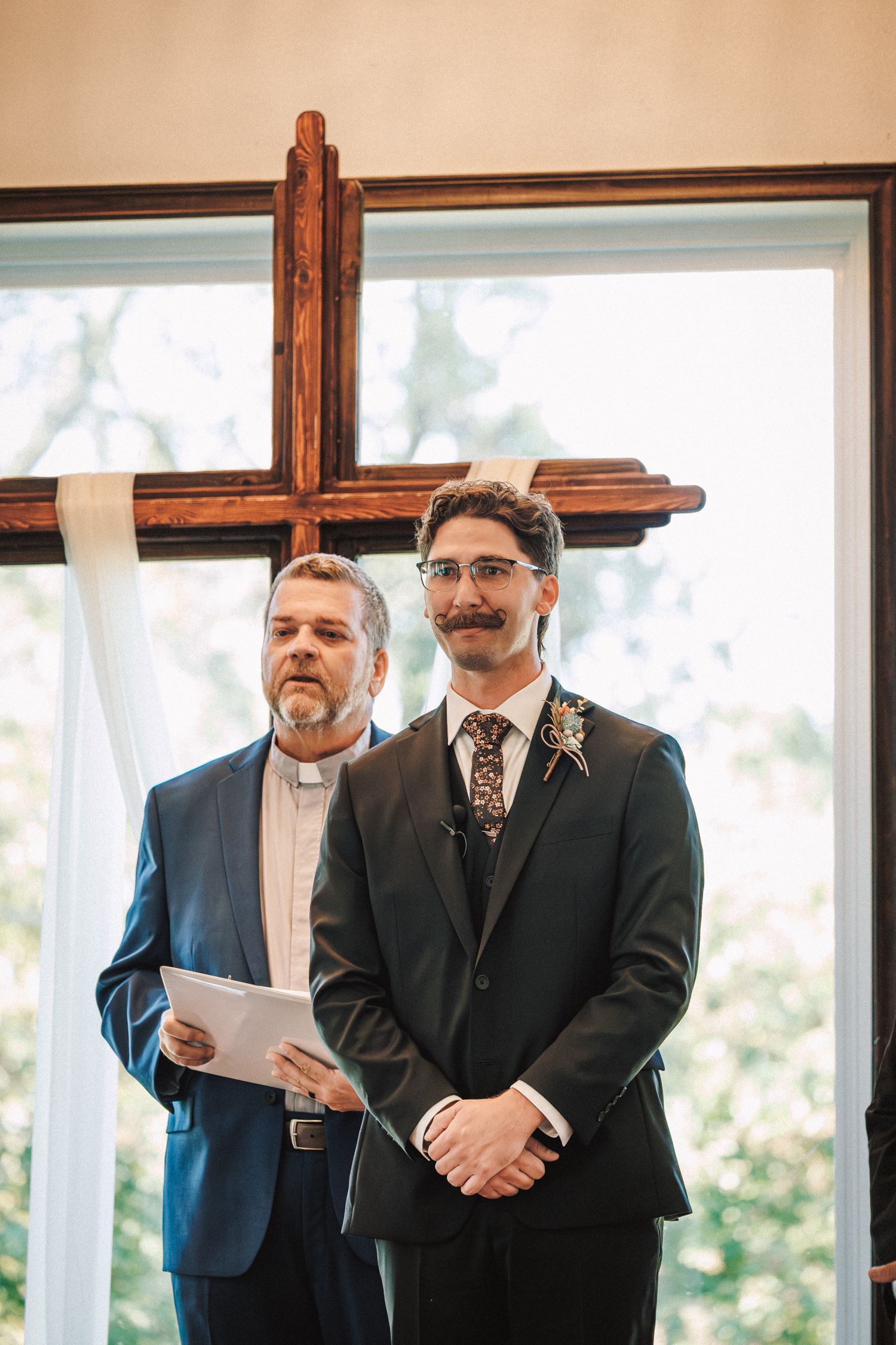 Groom wearing floral tie and boutonniere stands at altar with officiant before wooden cross in chapel.