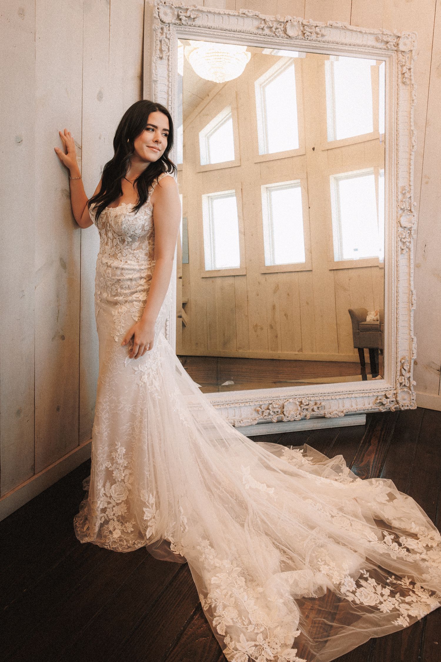 Bride in lace mermaid wedding gown with train poses beside ornate white mirror in rustic wood-paneled room.