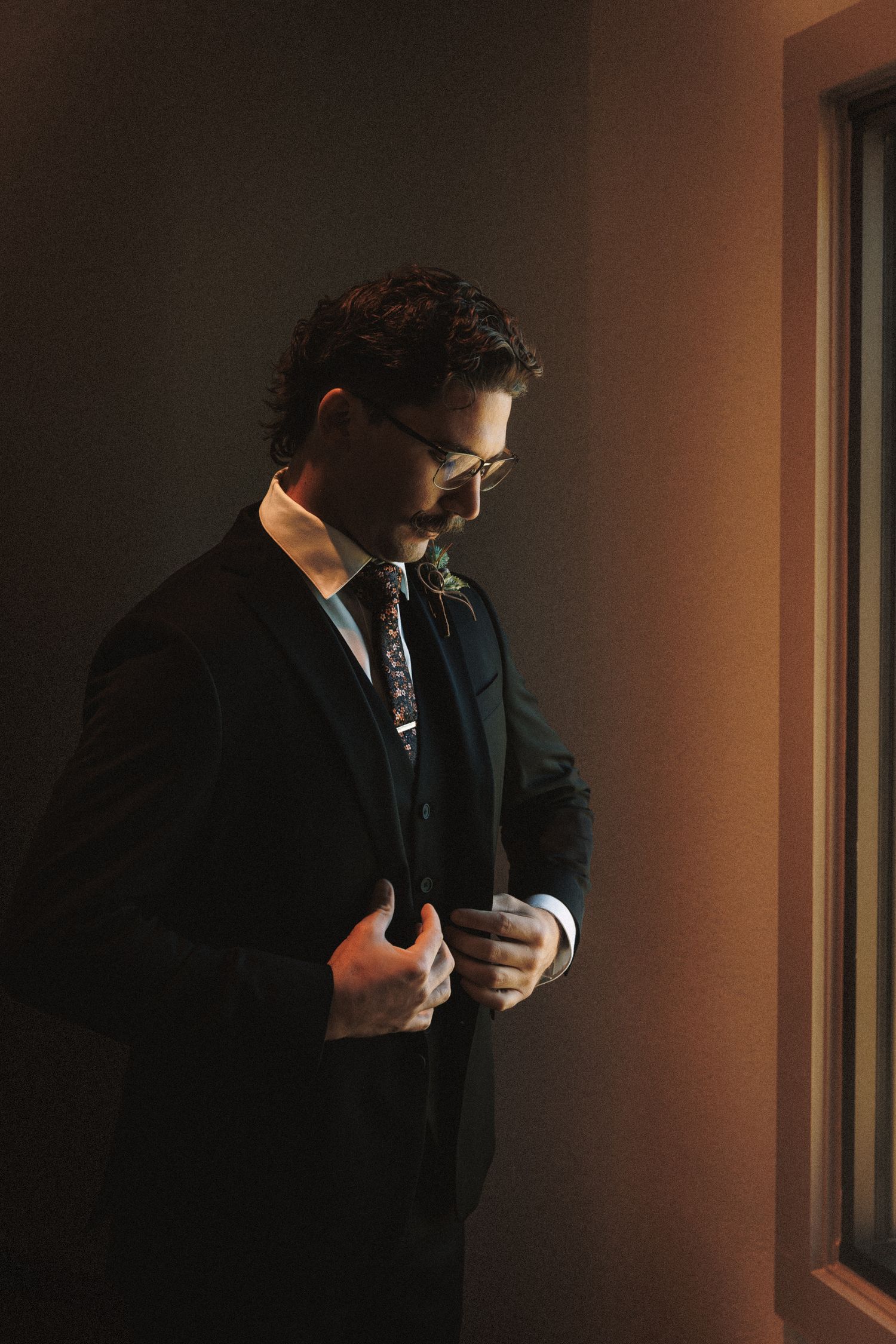 Dapper groom in dark suit adjusting jacket by window in moody natural light.