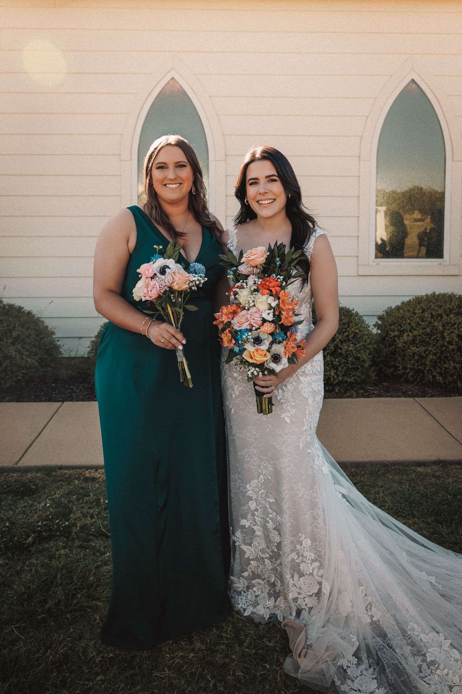 Bride in lace gown and bridesmaid in emerald dress holding bouquets outside white chapel.
