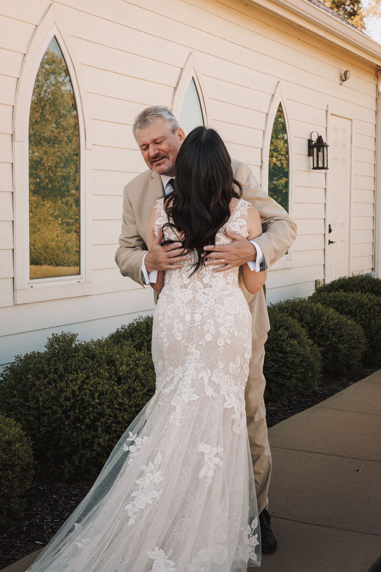 Emotional father embracing bride in lace mermaid gown outside white chapel wedding venue.
