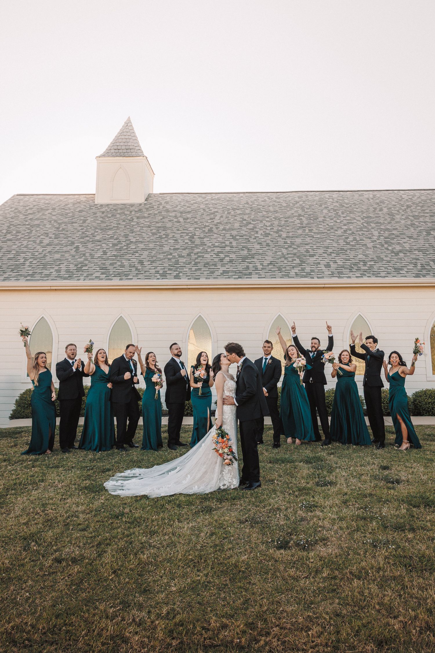Wedding party in teal dresses and black suits celebrate as bride and groom kiss outside a white chapel.