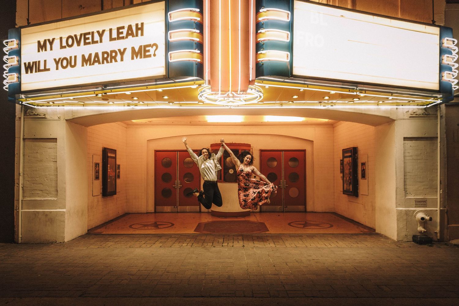 Couple jumping for joy beneath a bright theater marquee with a marriage proposal message.