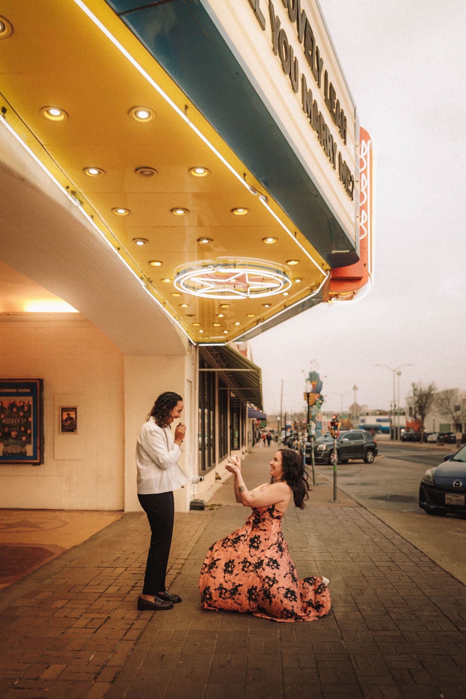 Couple dancing outside a vintage movie theater marquee with glowing yellow lights at dusk.