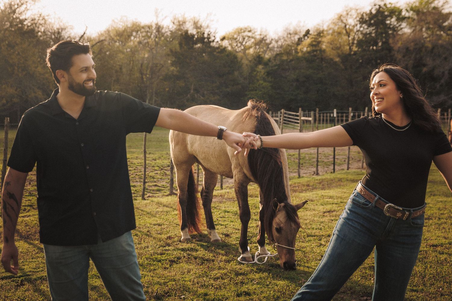 Couple holding hands and smiling with horses grazing in a sunlit farm field.