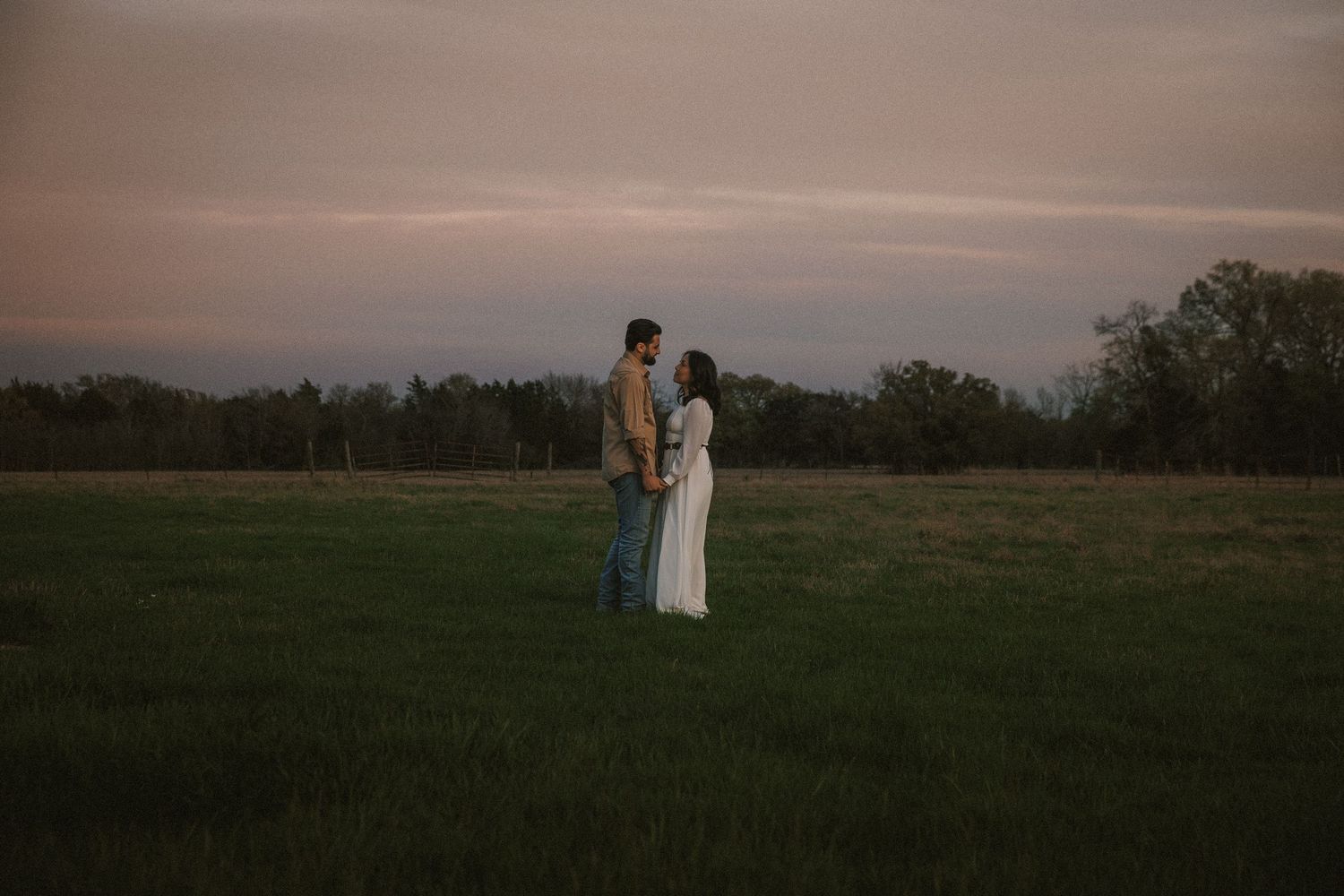 Couple standing face to face holding hands in a vast green field at dusk.