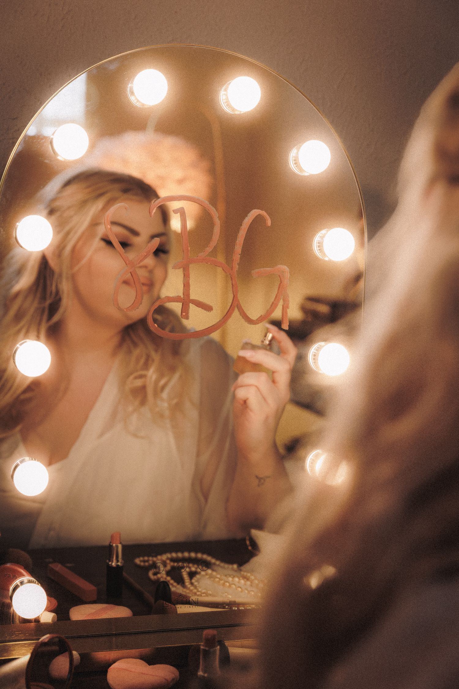 Blonde bride holding monogram sign reflected in Hollywood vanity mirror with glowing bulbs during bridal prep.