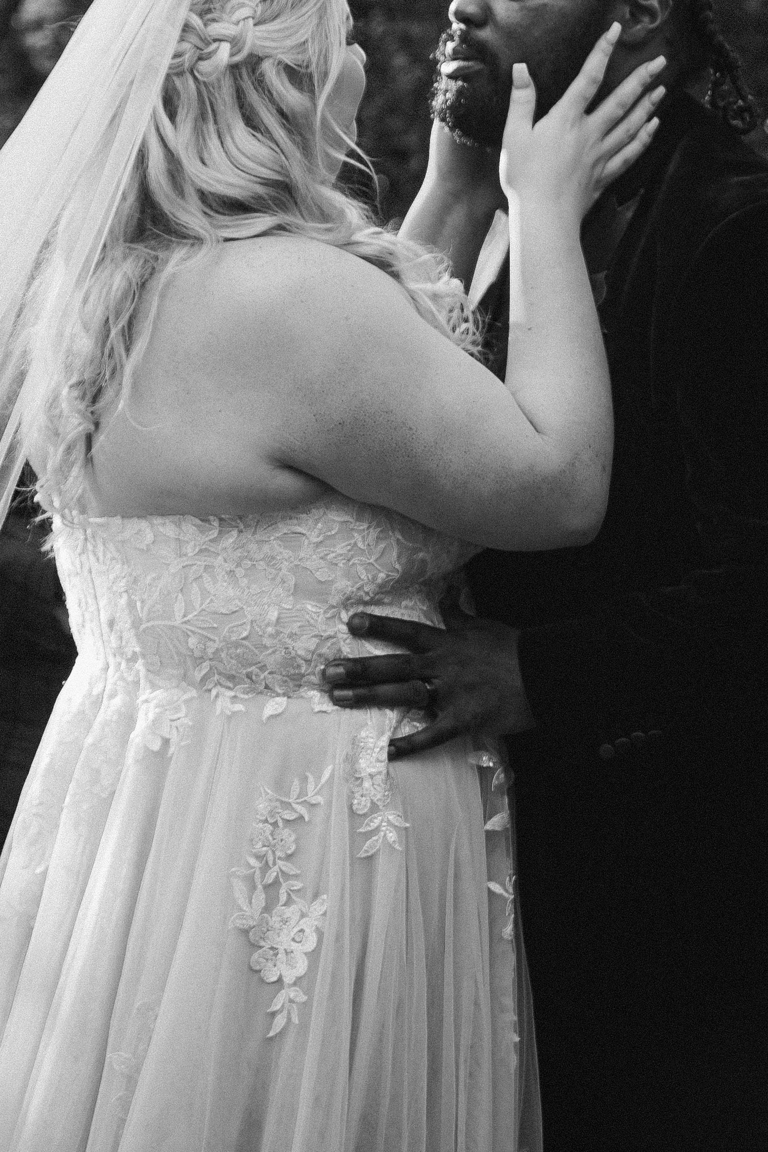 Black and white photo of bride and groom embracing, showcasing lace wedding gown detail and tender moment.