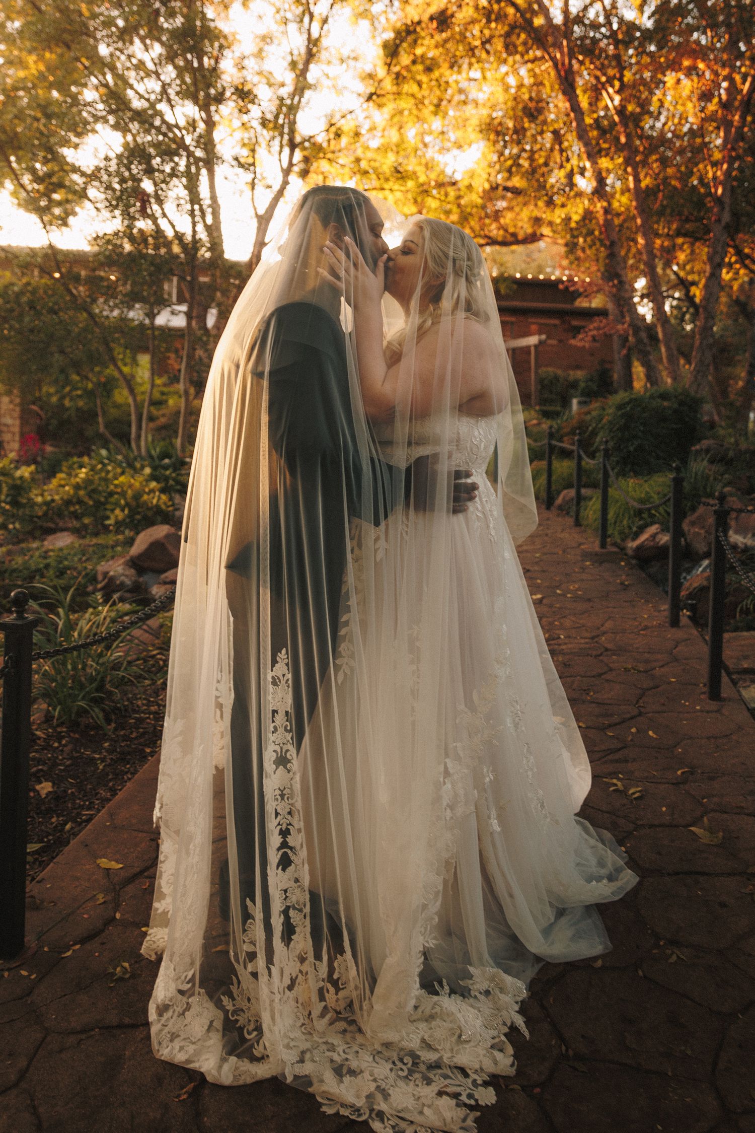 Bride and groom kissing under a long lace-trimmed veil on a garden pathway at golden hour sunset.