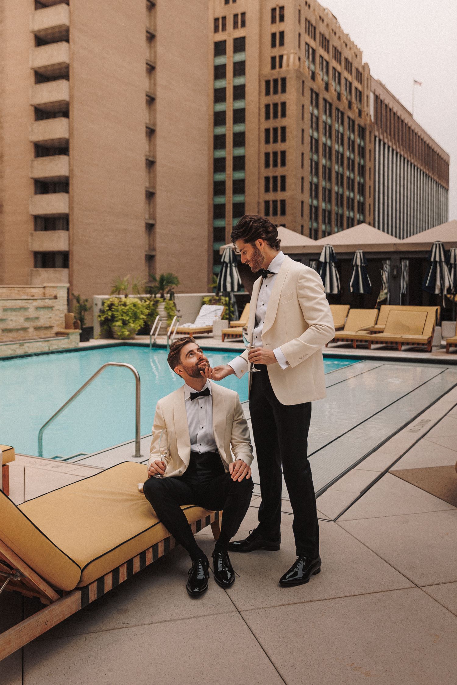 Two grooms in cream tuxedo jackets sharing a moment by rooftop hotel pool.