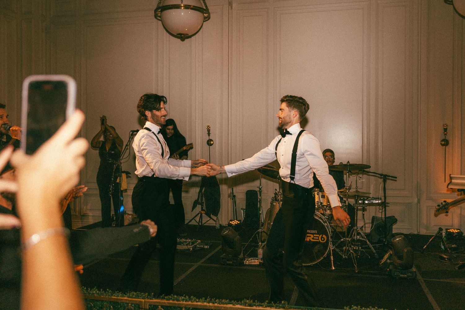 Two men in white shirts and suspenders dancing joyfully at wedding reception with live band in background.