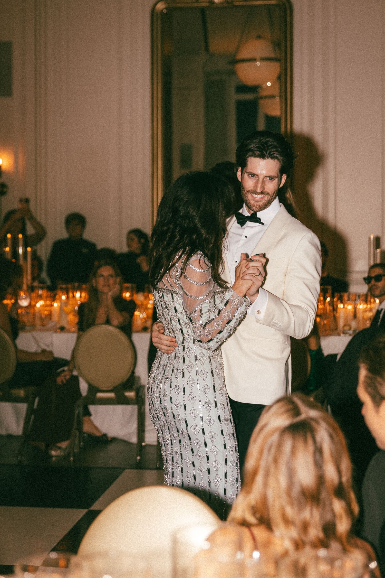 Couple sharing first dance at elegant wedding reception, groom in white tuxedo jacket holding bride in silver beaded gown.