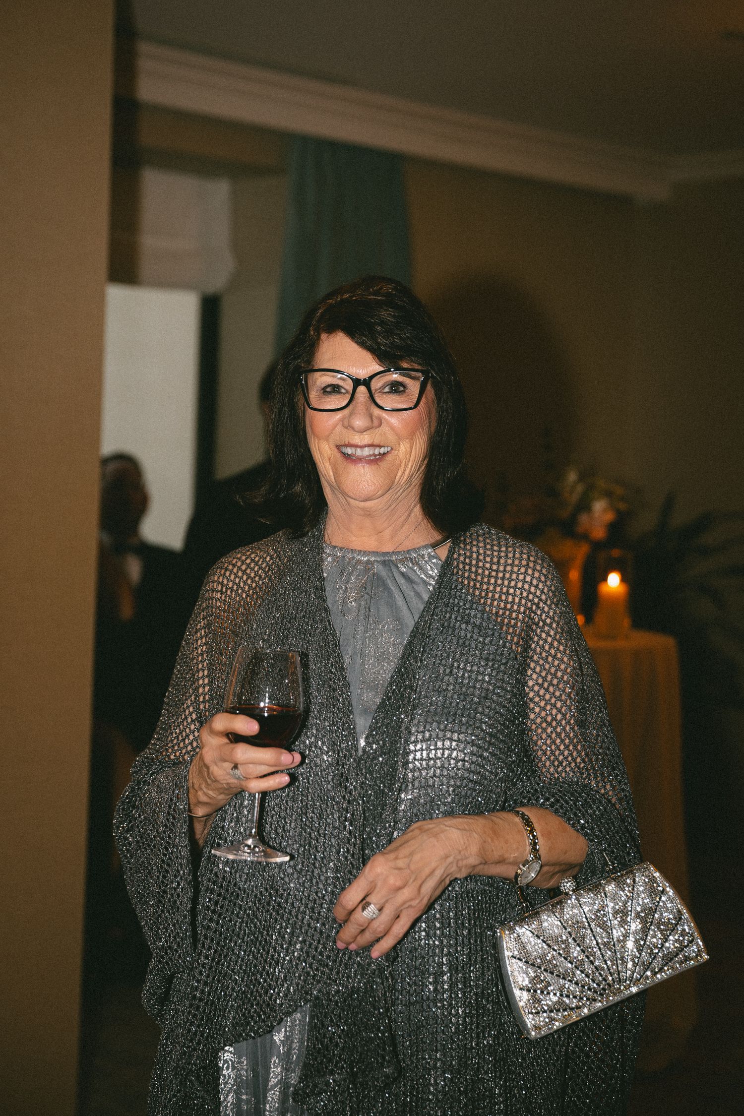 Elegant older woman in silver sequined dress holding a glass of red wine and a metallic clutch at a wedding.
