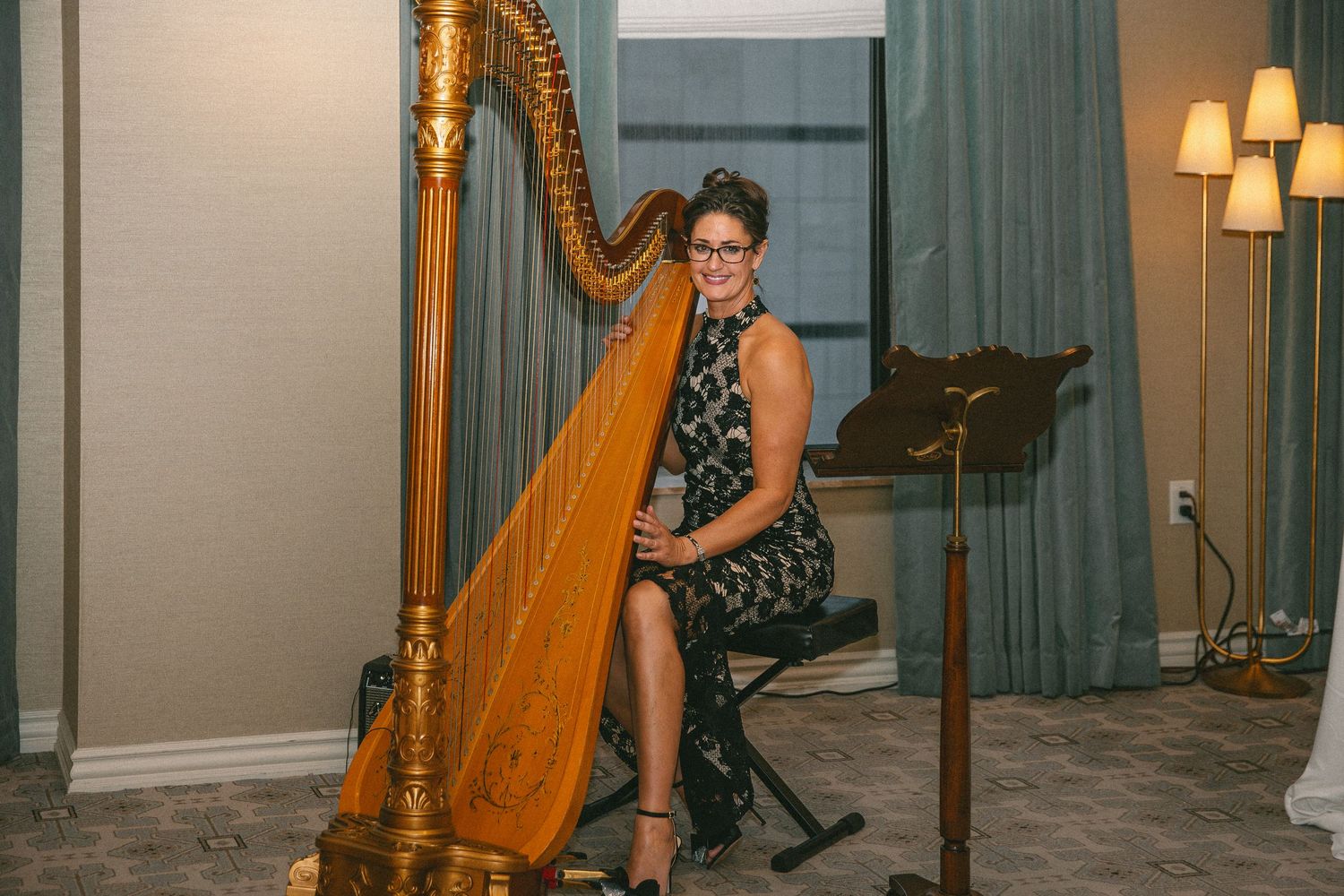 A woman in a floral dress smiles while playing a golden harp at a wedding.