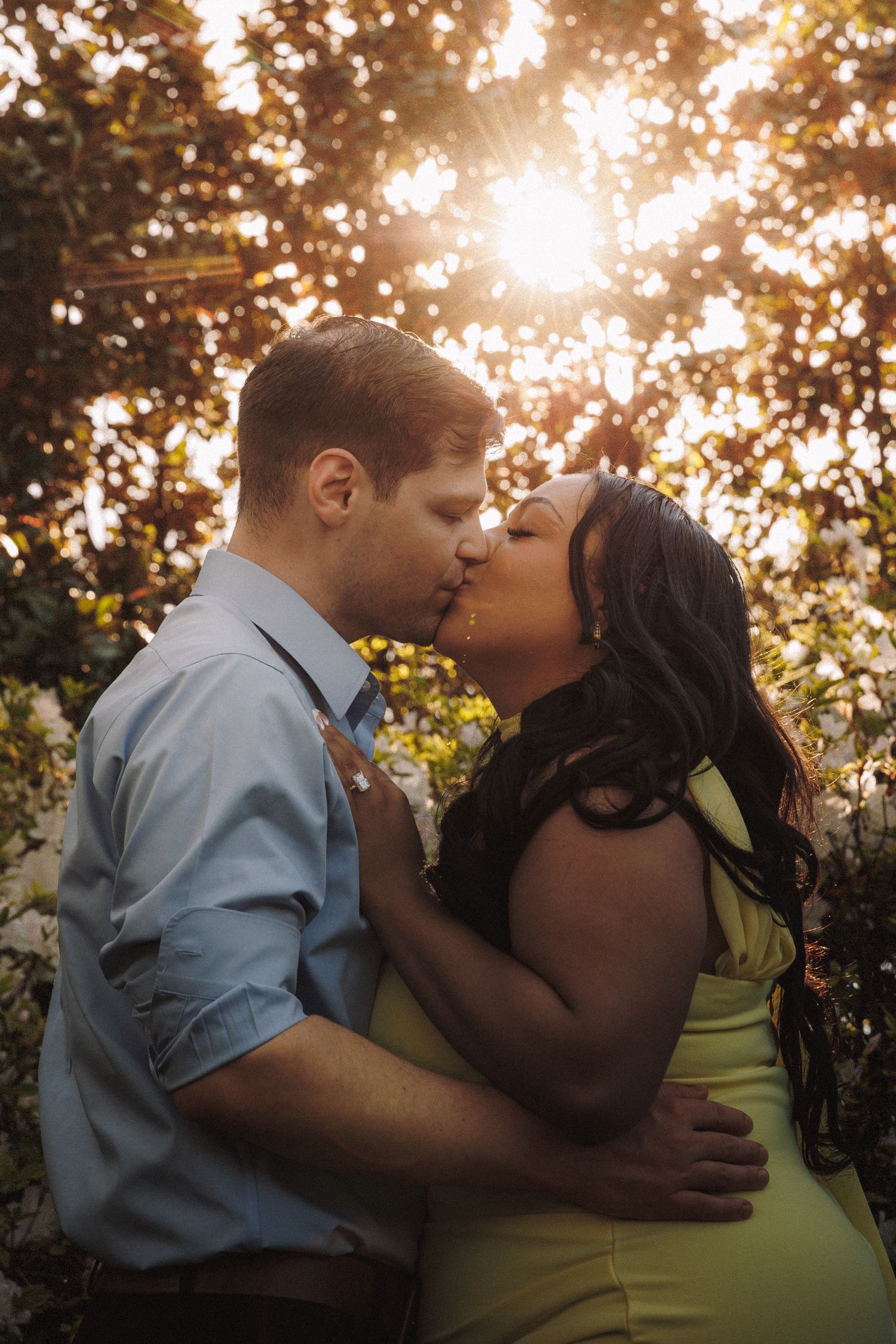 Couple kissing in front of sun-drenched flowering bushes, woman in green dress and man in blue shirt in warm light.