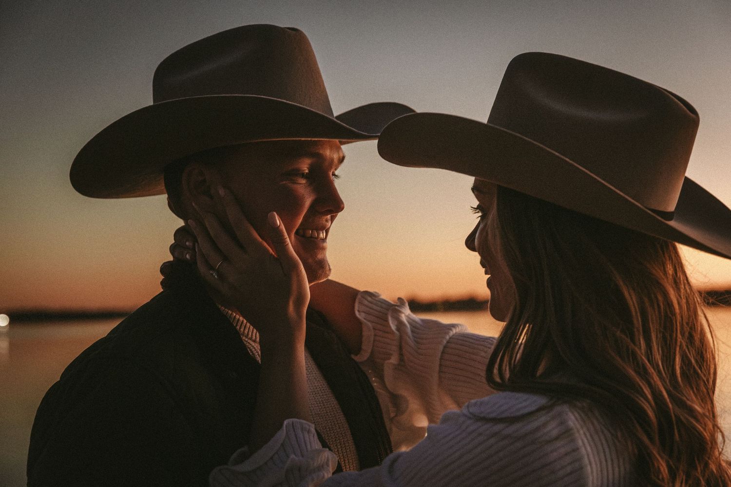 Romantic couple in cowboy hats share an intimate moment against a golden sunset backdrop.
