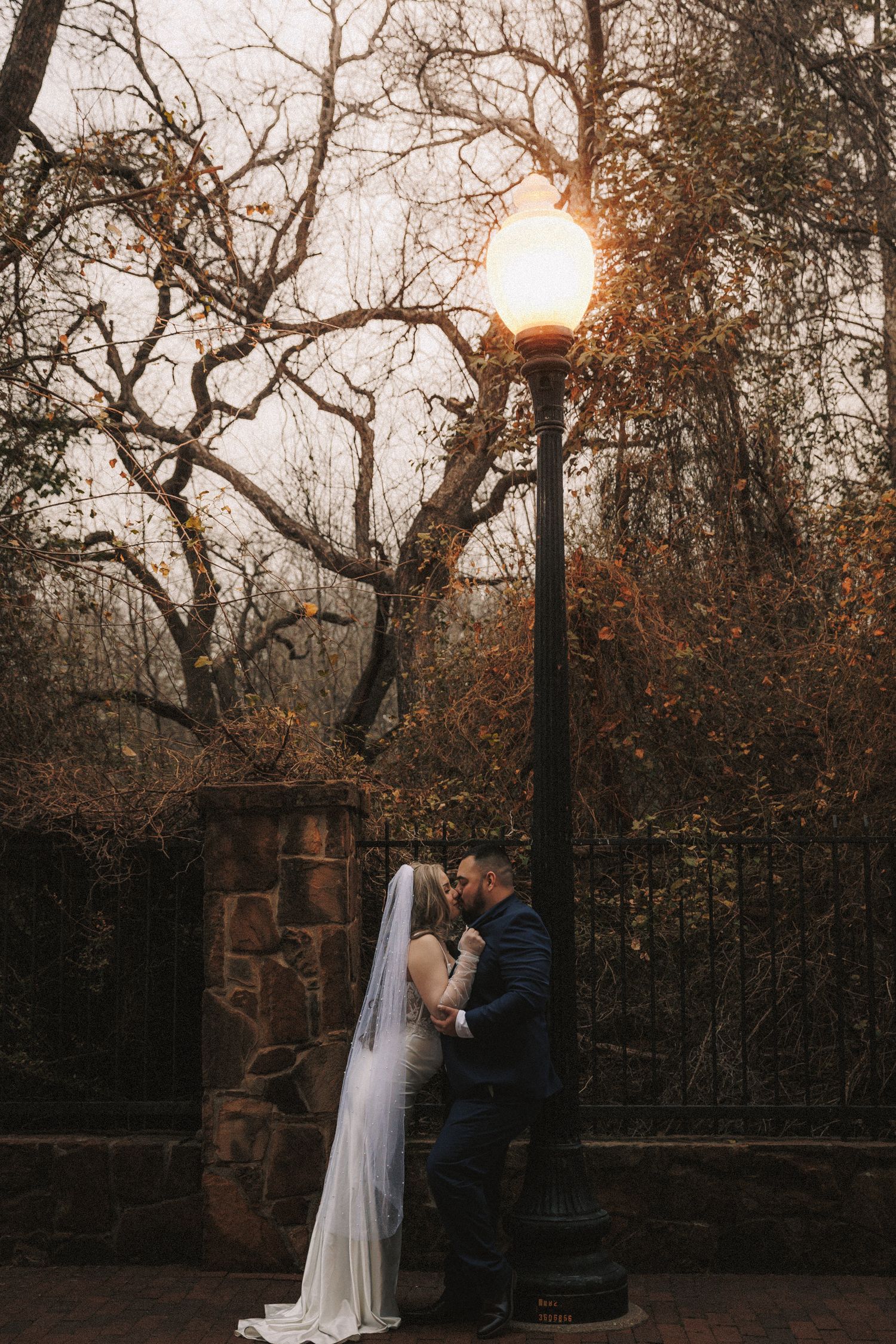 A romantic couple shares an intimate moment under a glowing lamp post at sunset in a park setting.