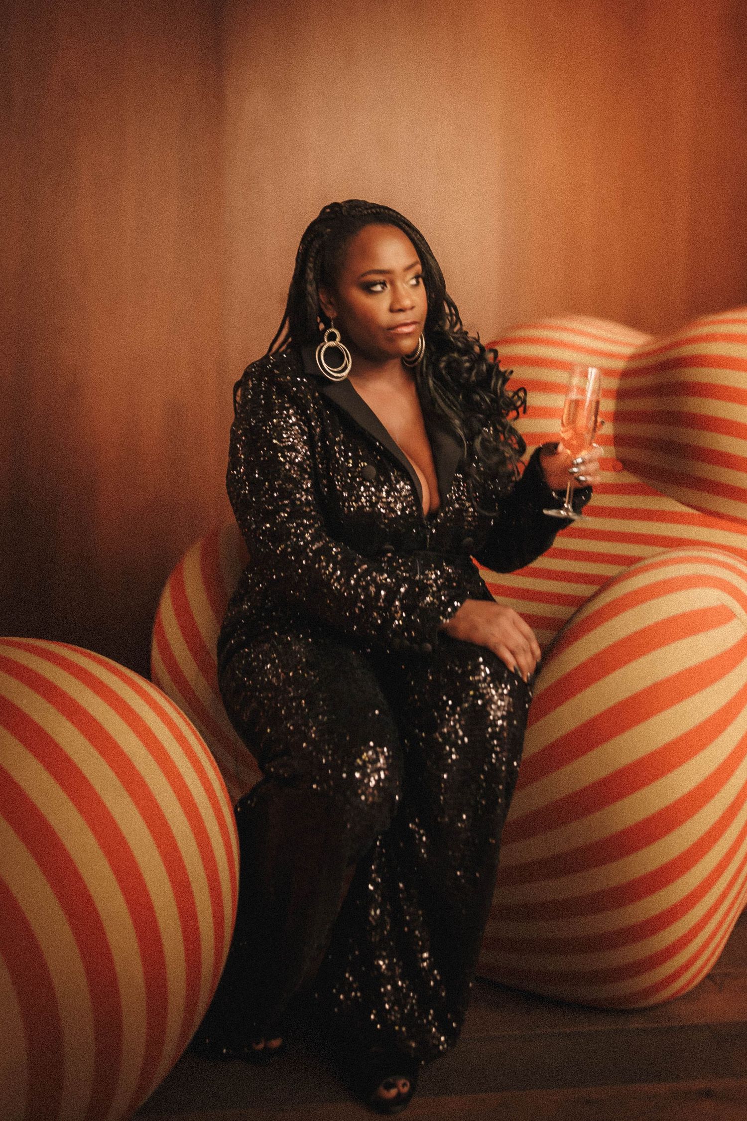 A person in a sparkly black evening gown poses with a champagne glass against orange striped spherical backdrop.
