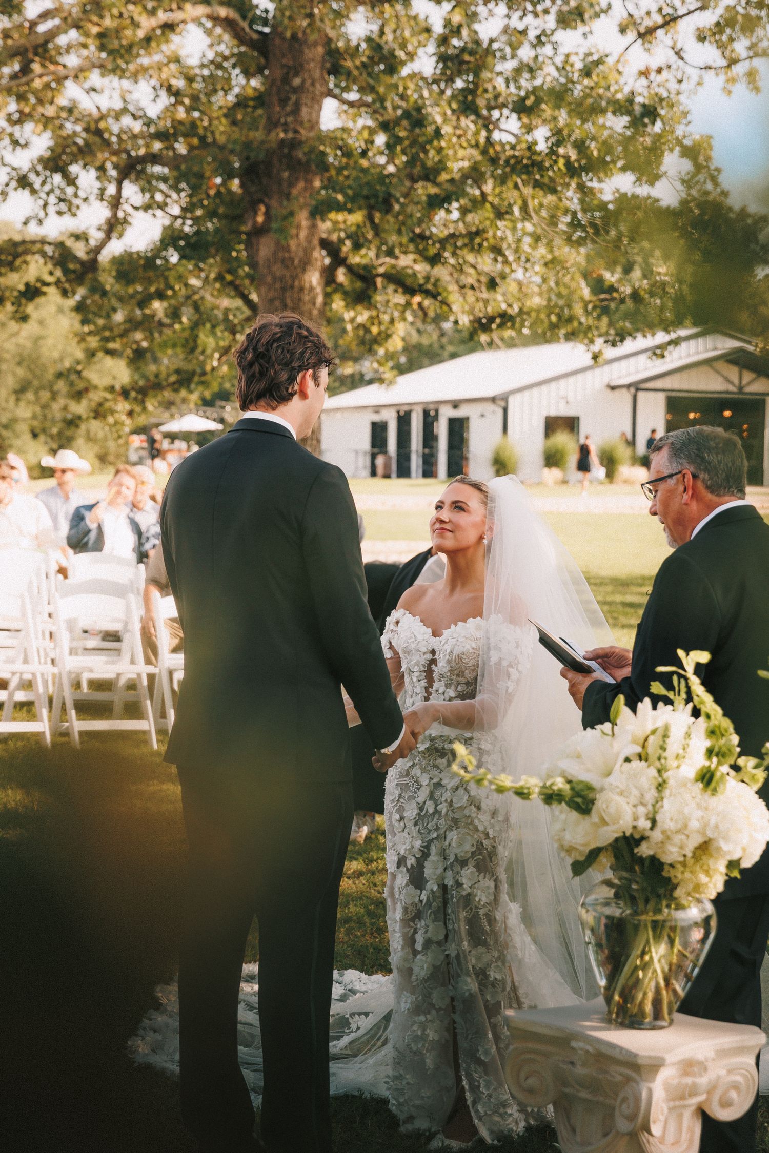 Wedding ceremony group stands along an outdoor walkway with white floral arrangements and chairs.