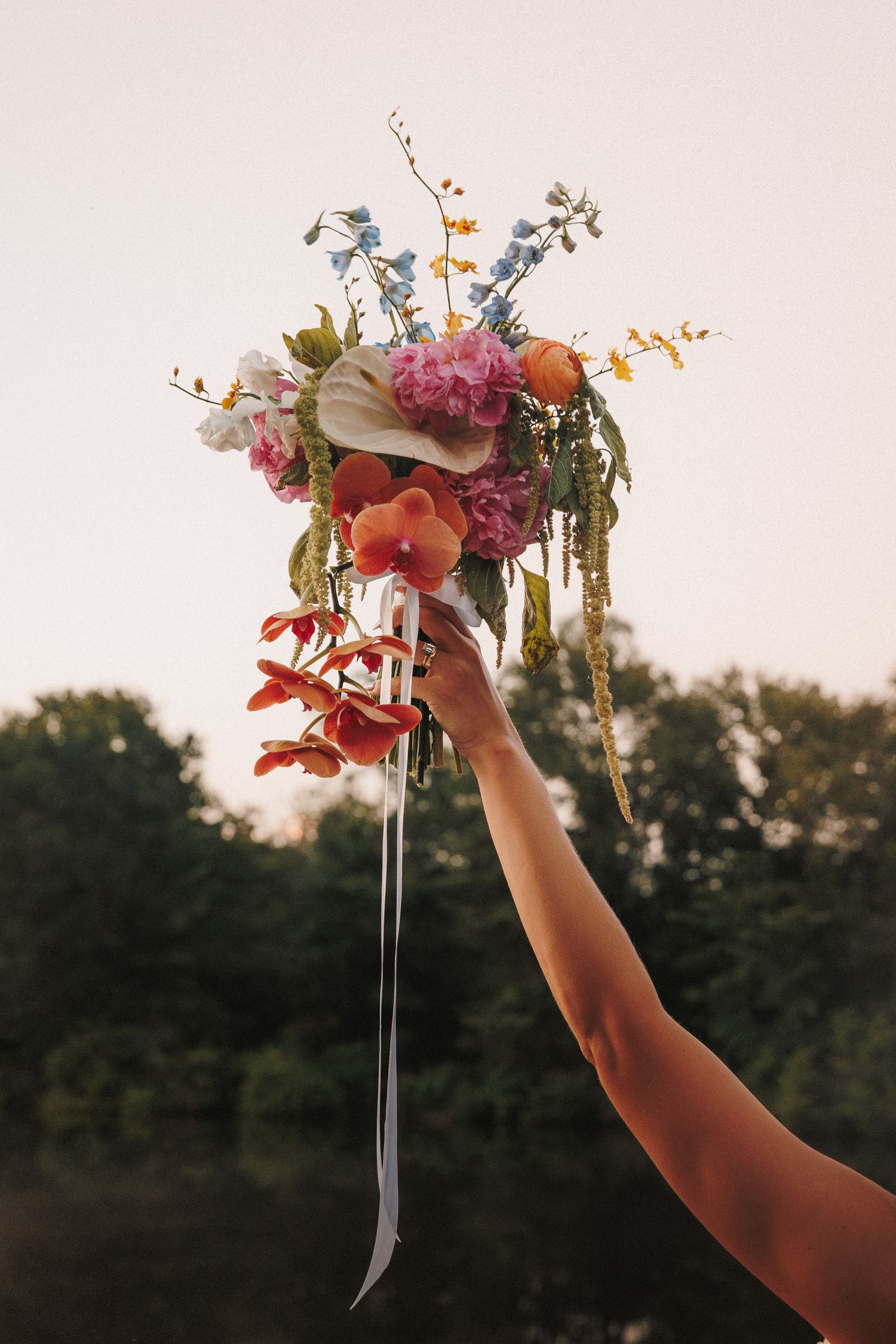 A hand holds up a floral bouquet arrangement against a sunset background in nature.