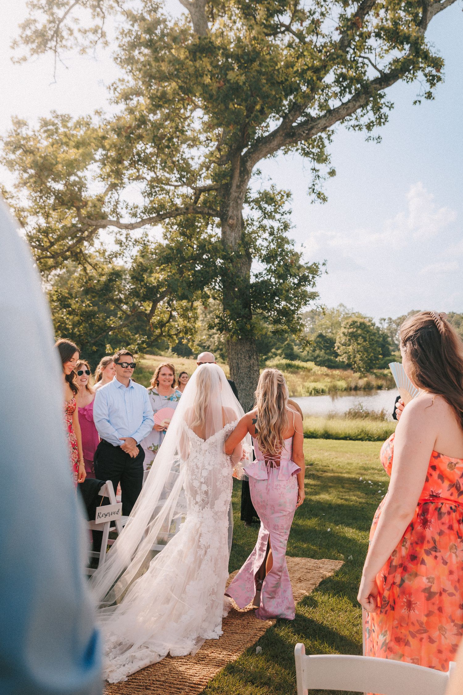 Wedding party walking together across sunny lawn in floral dresses and gowns during outdoor summer celebration.