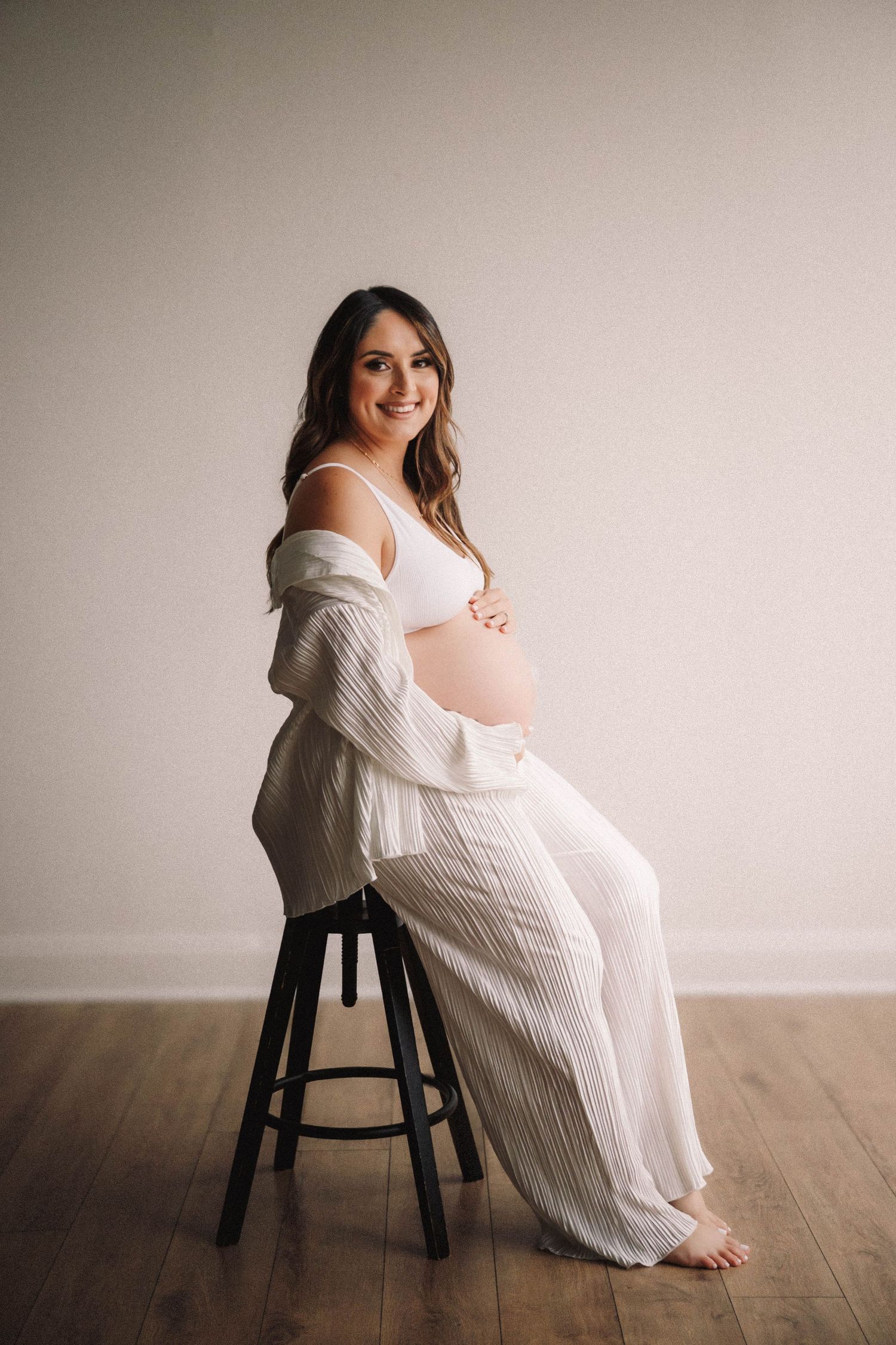 A series of maternity photos showing someone in a white sweater dress sitting on a dark wooden stool against a neutral wall.