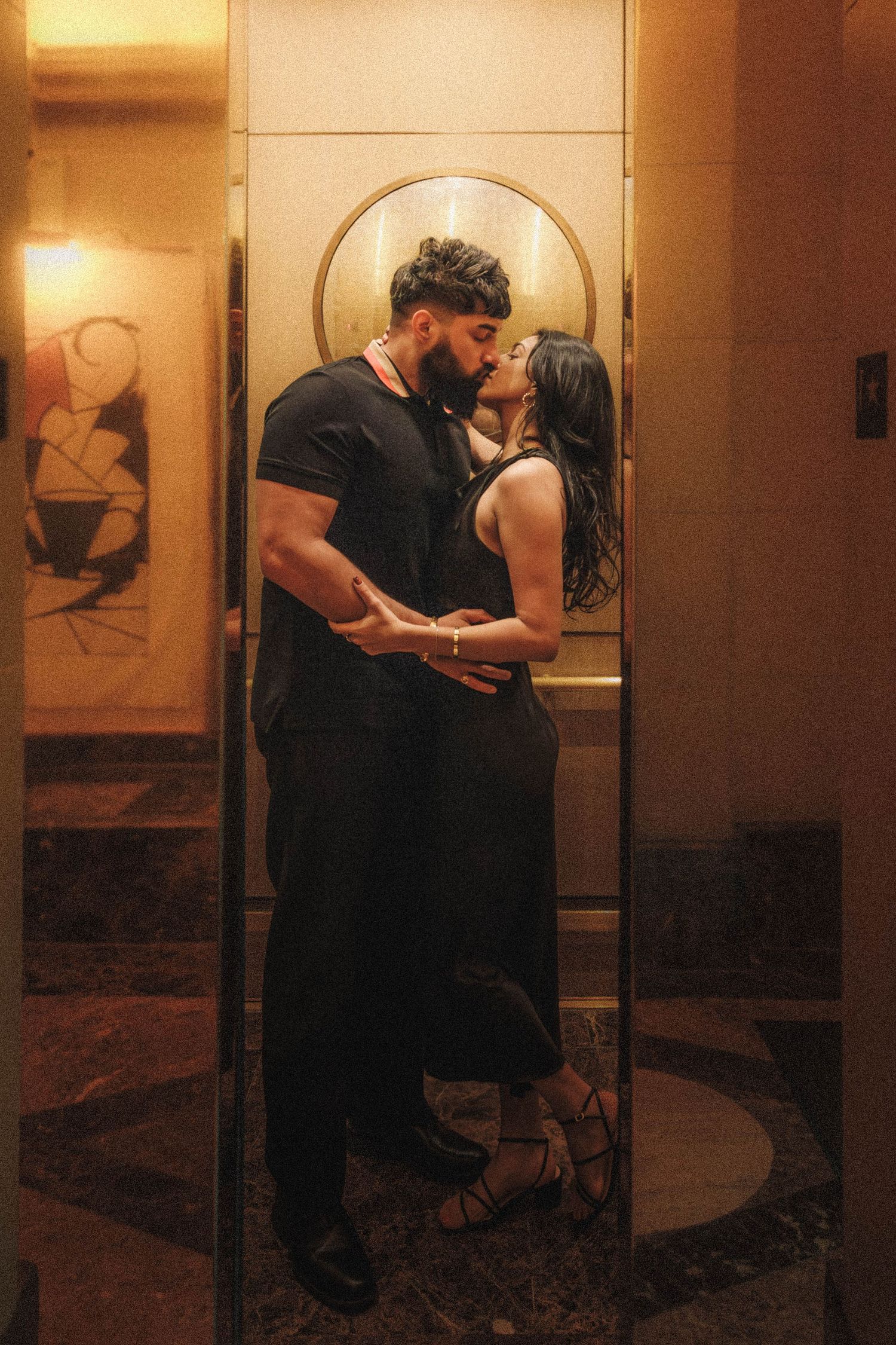 A couple embraces in a warmly lit interior hallway with artistic wall decorations.