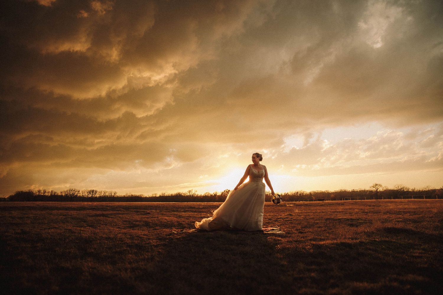A bride stands in silhouette against a dramatic sunset sky with golden clouds over an open field.