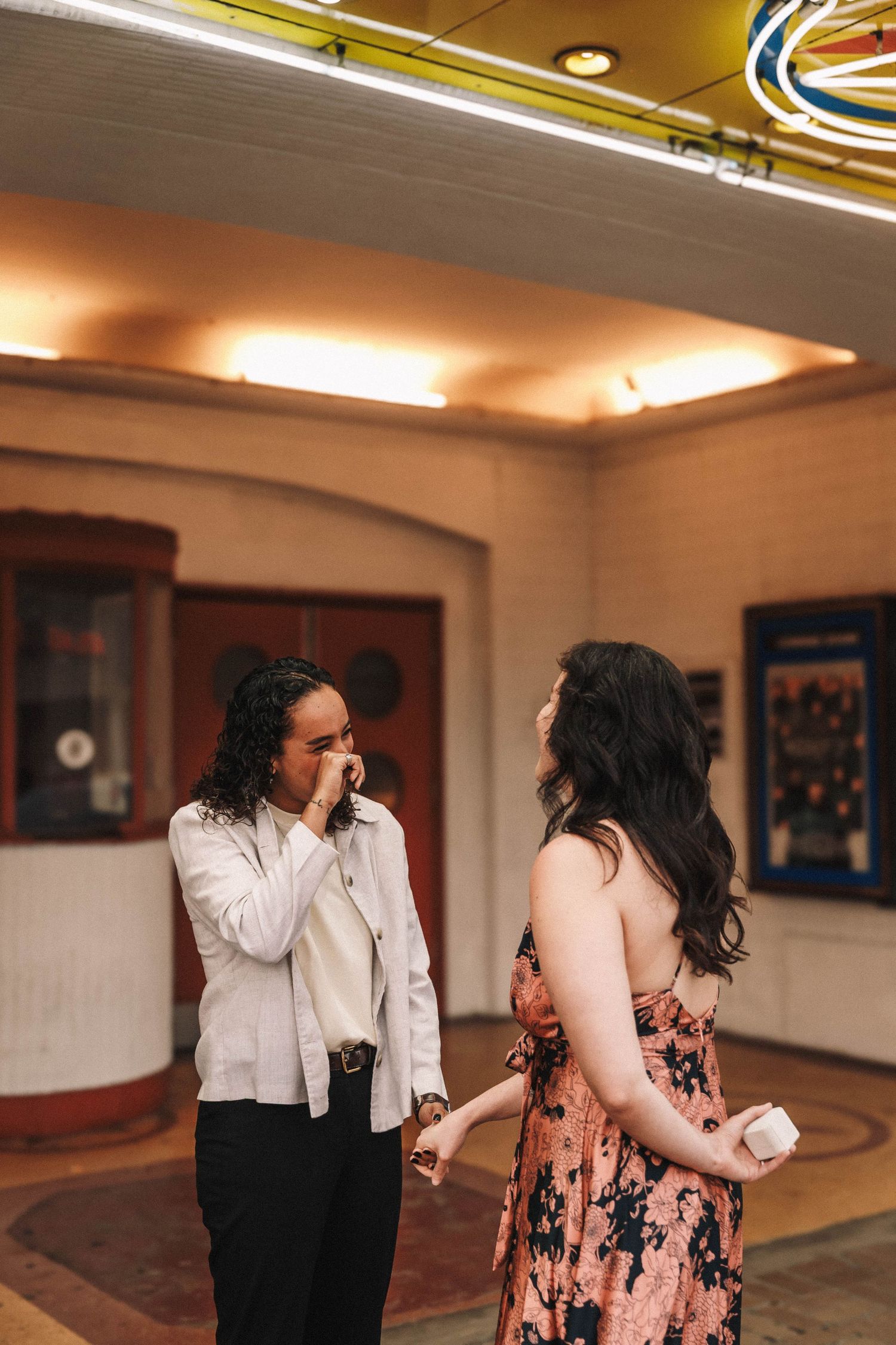 Two women share an emotional moment inside a vintage theater lobby with neon lights.
