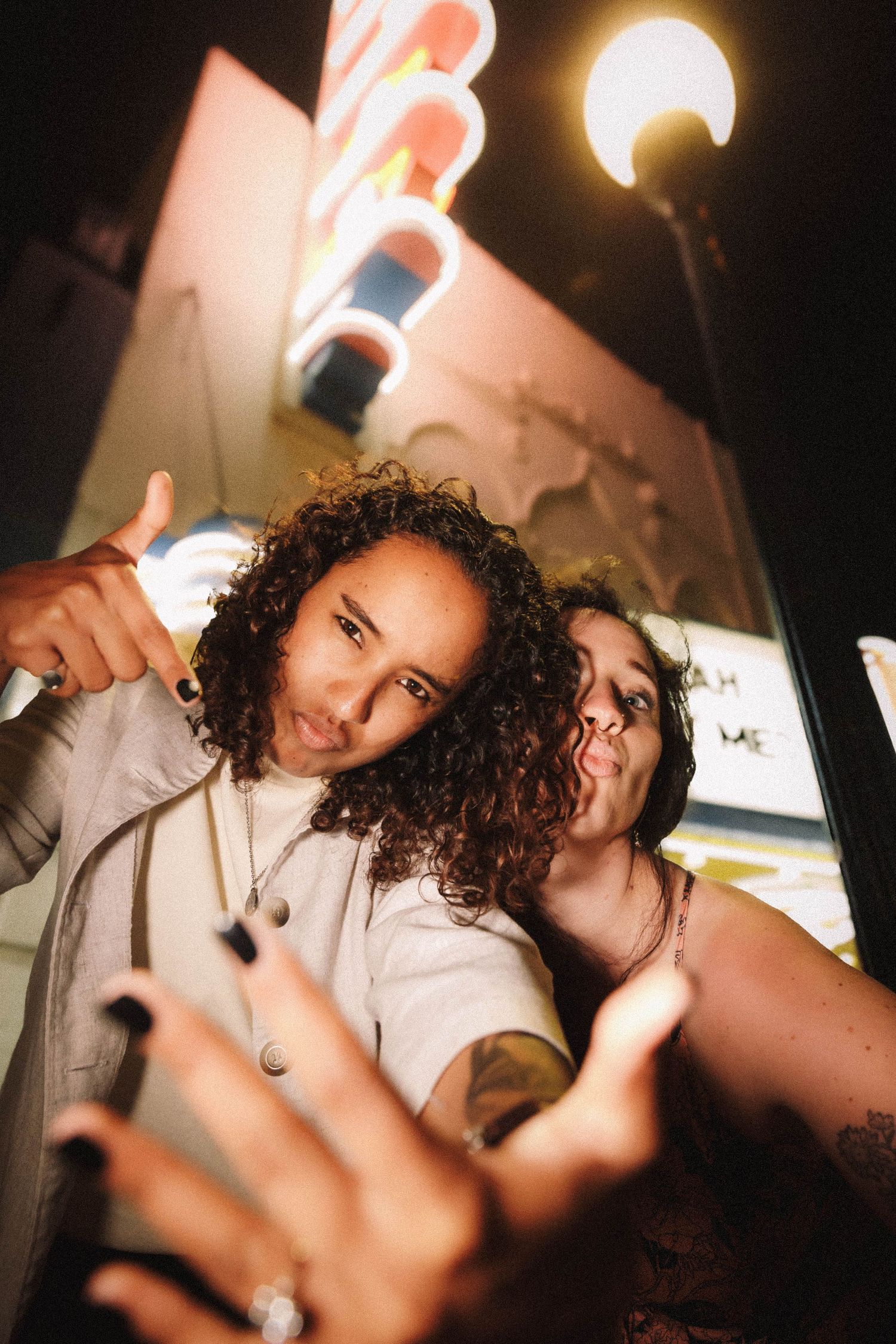 Two women playfully pose for a selfie under neon lights outside a theater at night.