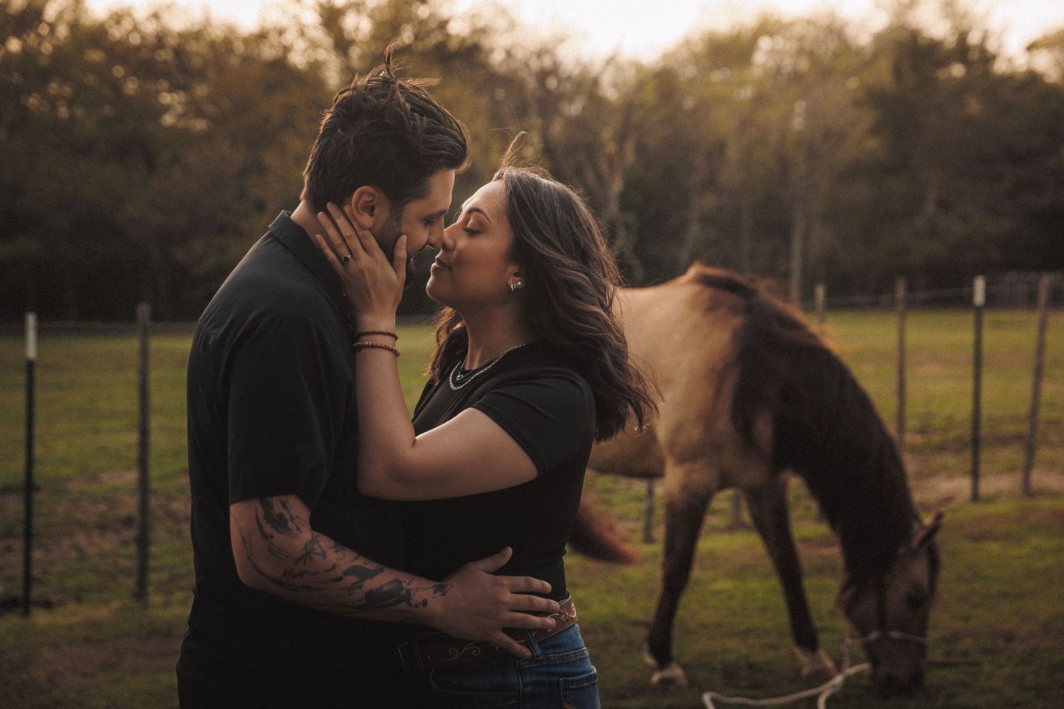 Couple sharing a romantic almost-kiss at golden hour on a farm with a horse grazing in the background.