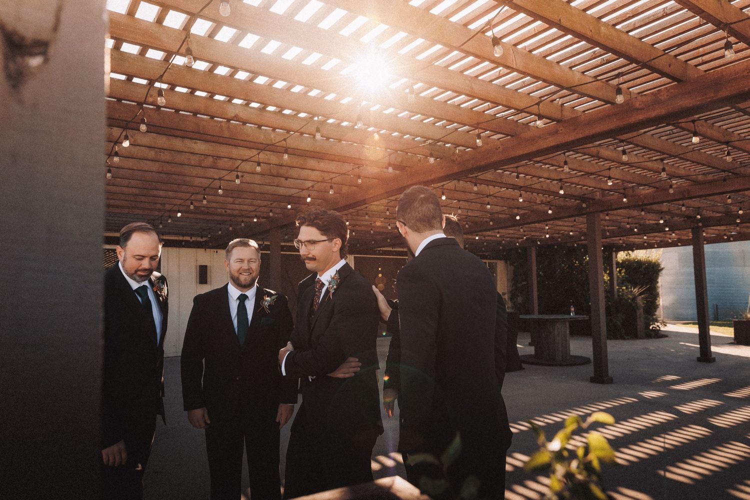 Groomsmen in suits gather under a wooden pergola with string lights in golden hour sunlight.