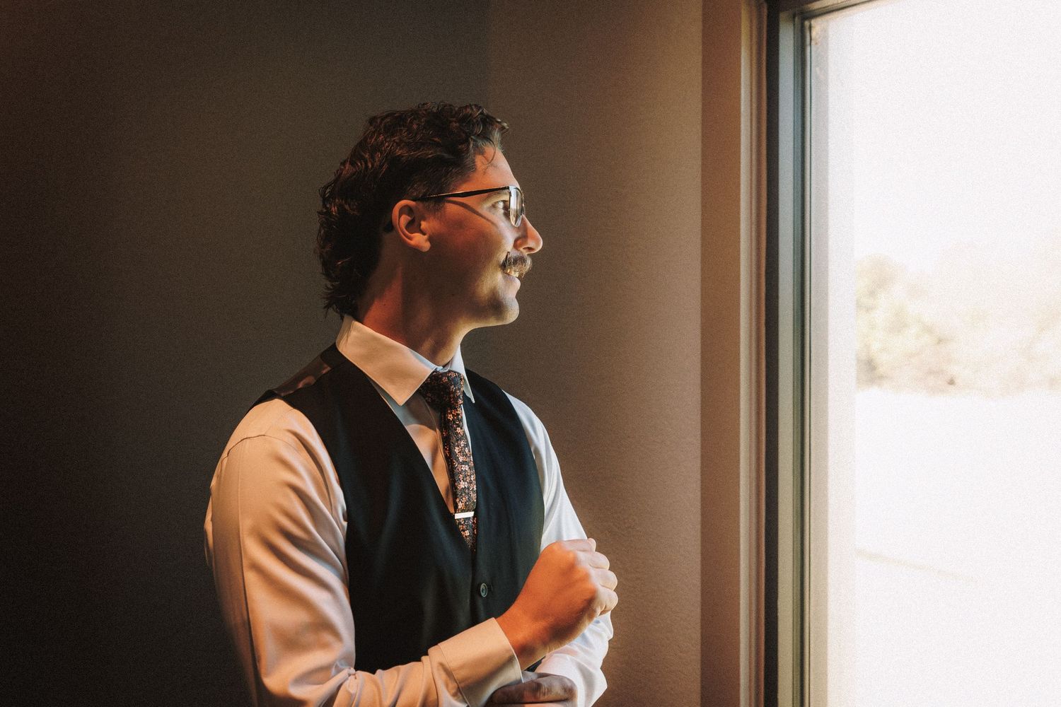Groom in vest and floral tie gazes thoughtfully out window in soft natural light.