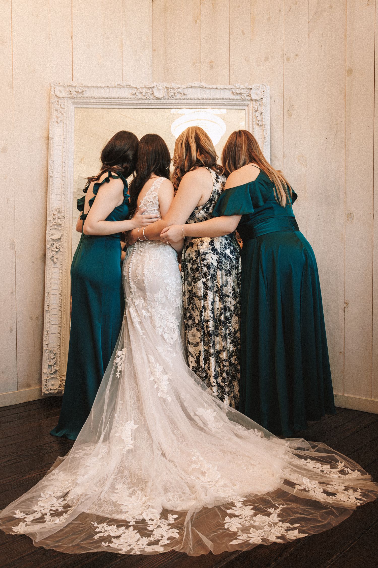 Bride in lace gown embraced by bridesmaids in teal dresses facing a mirror.