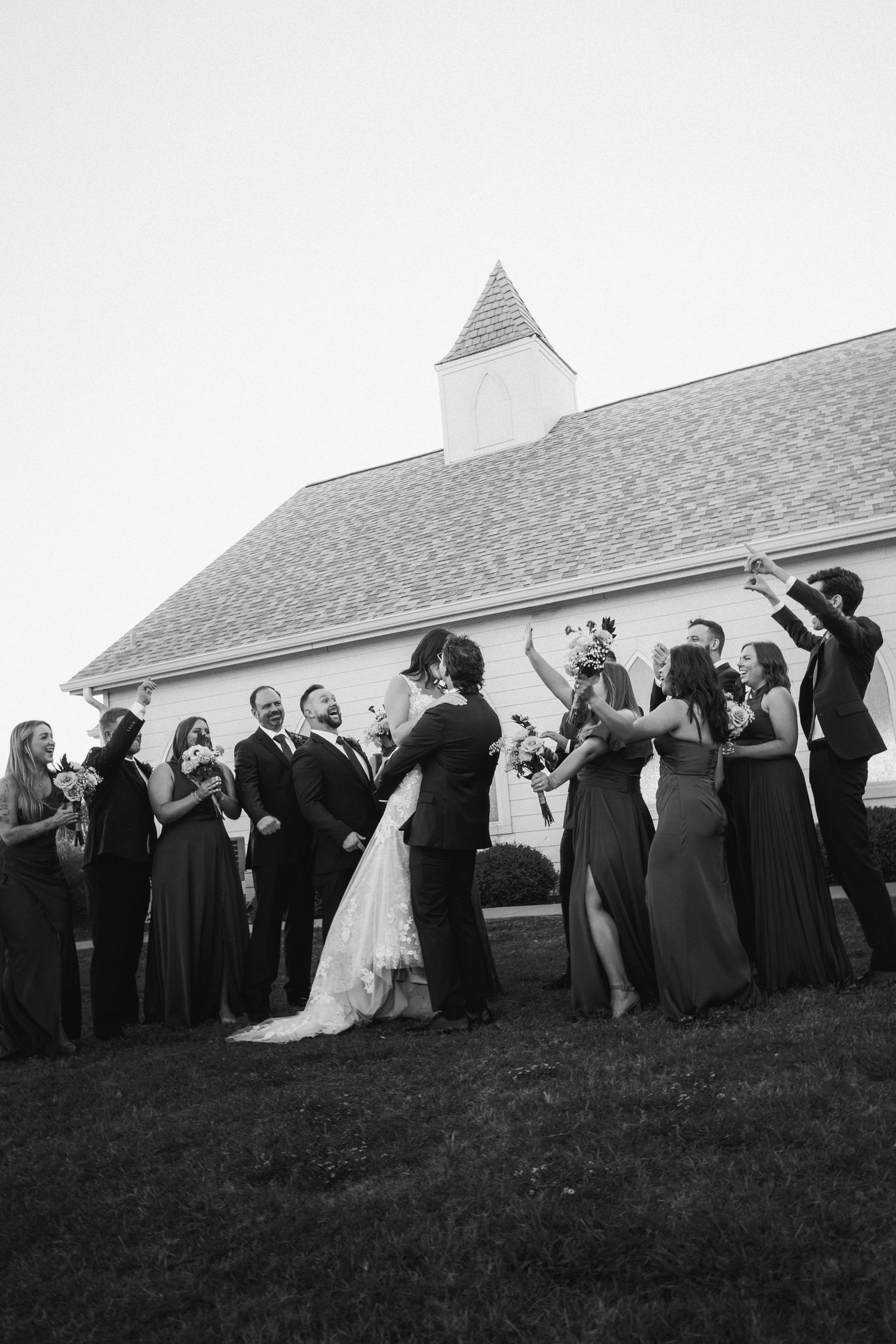 Black and white photo of wedding party celebrating outside white chapel after ceremony.
