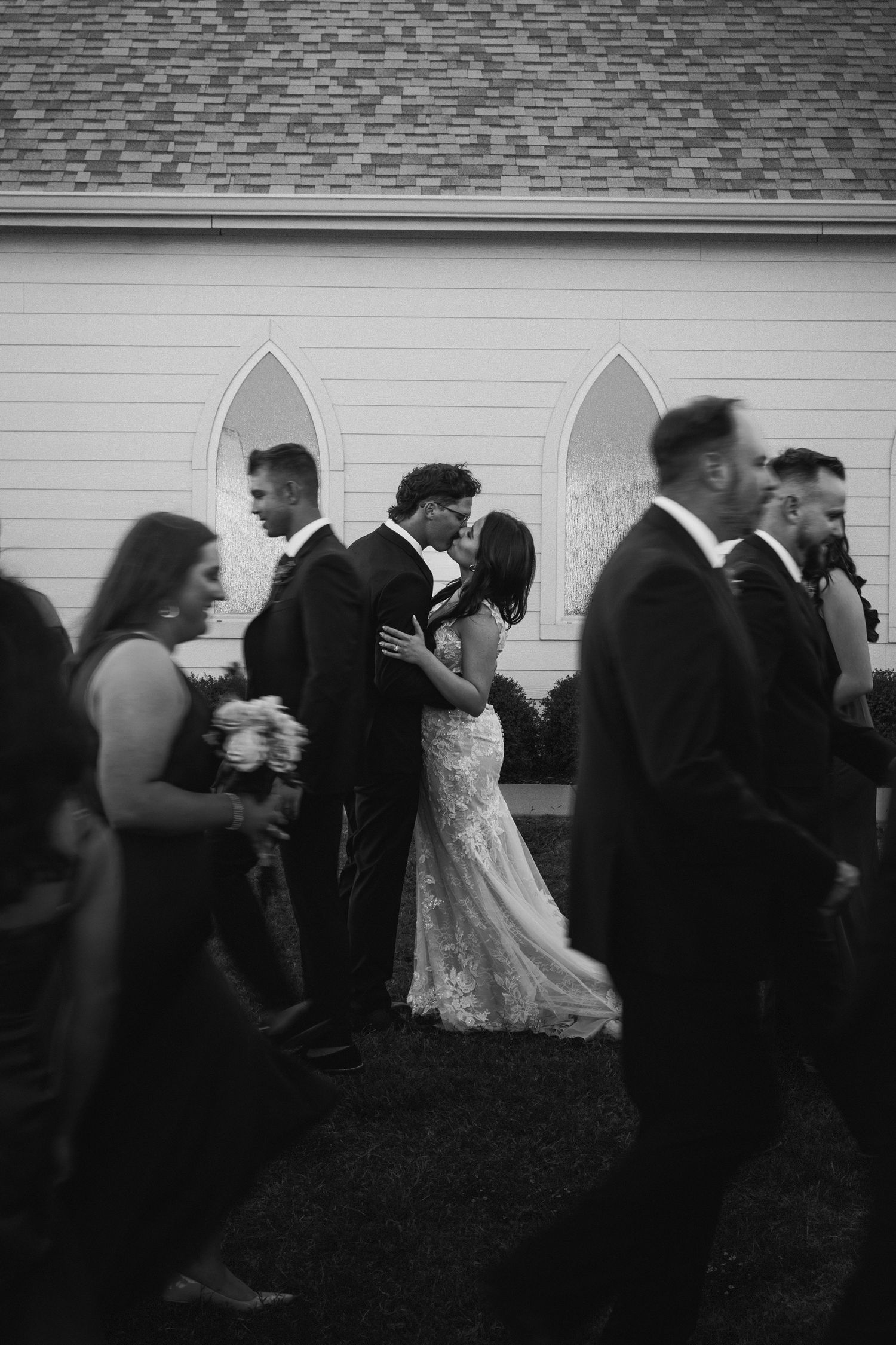 Bride and groom share a first kiss outside a white chapel surrounded by wedding party in black and white photo.