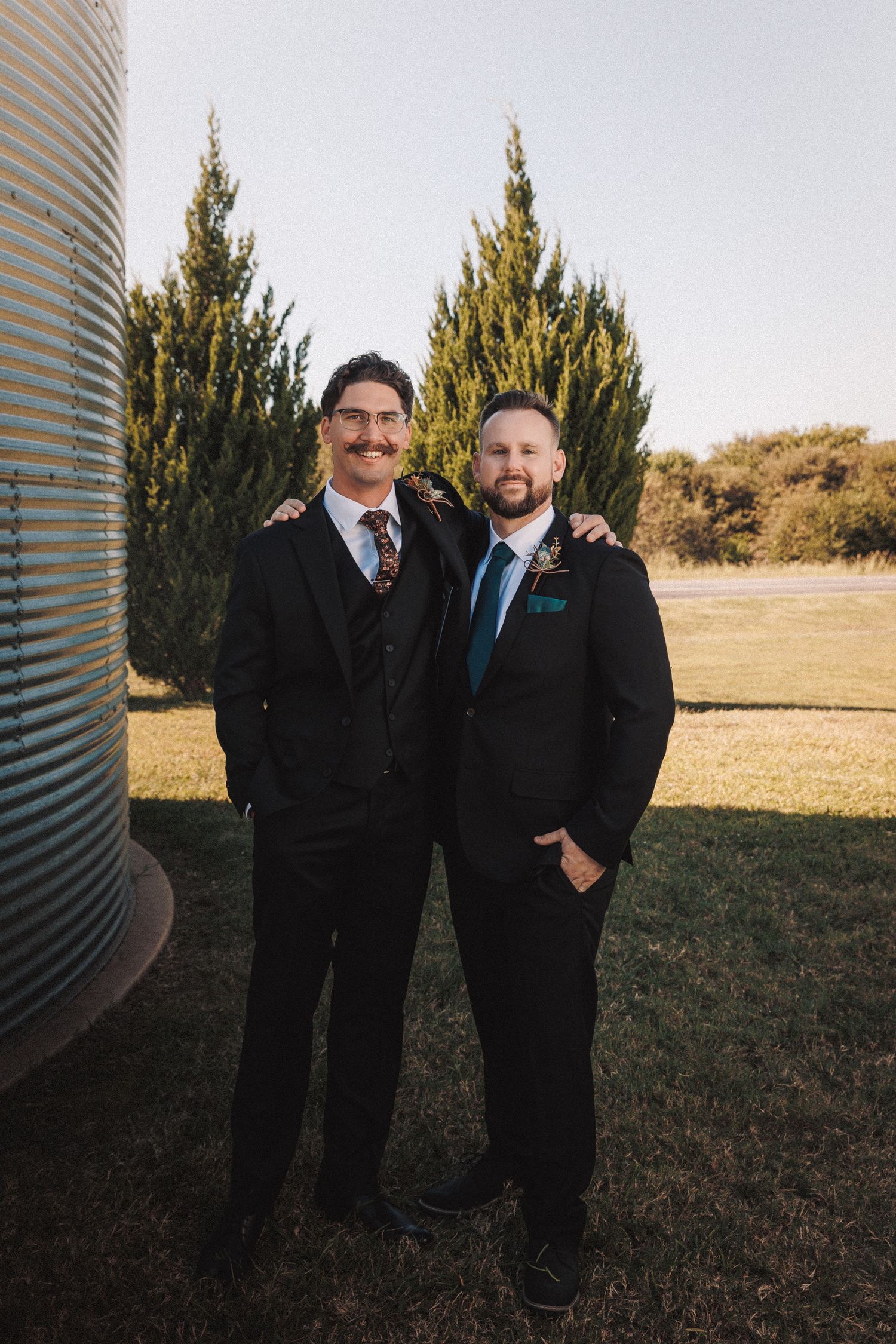 Two groomsmen in black suits smile together outdoors near grain silo and evergreen trees at wedding venue.