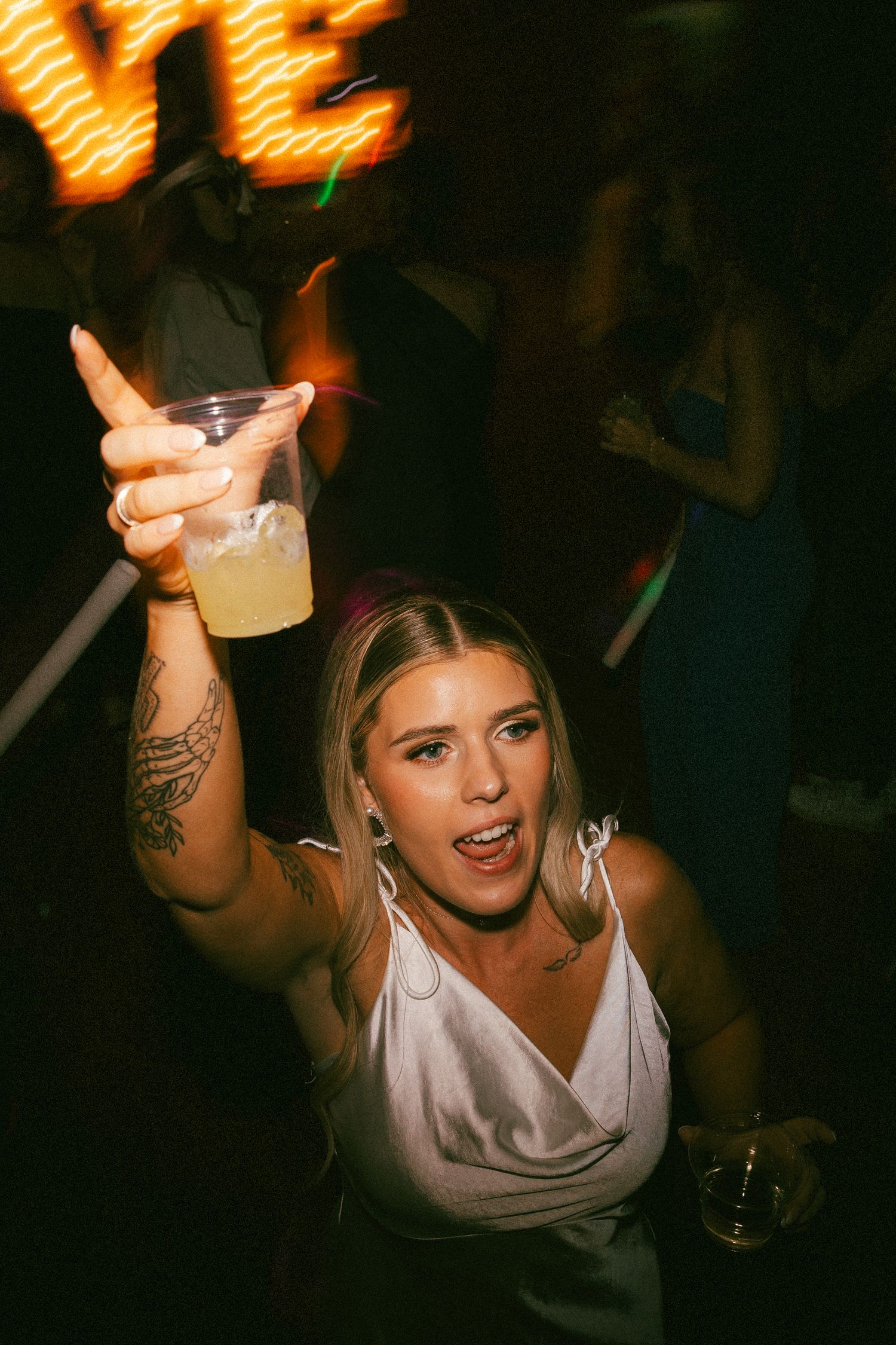A blonde woman in white raises a drink celebrating at a lively wedding reception.