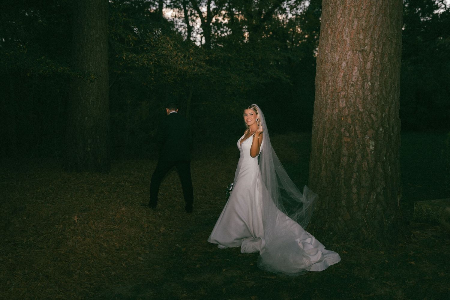 Bride in flowing white gown and veil walking with groom among tall trees at dusk.
