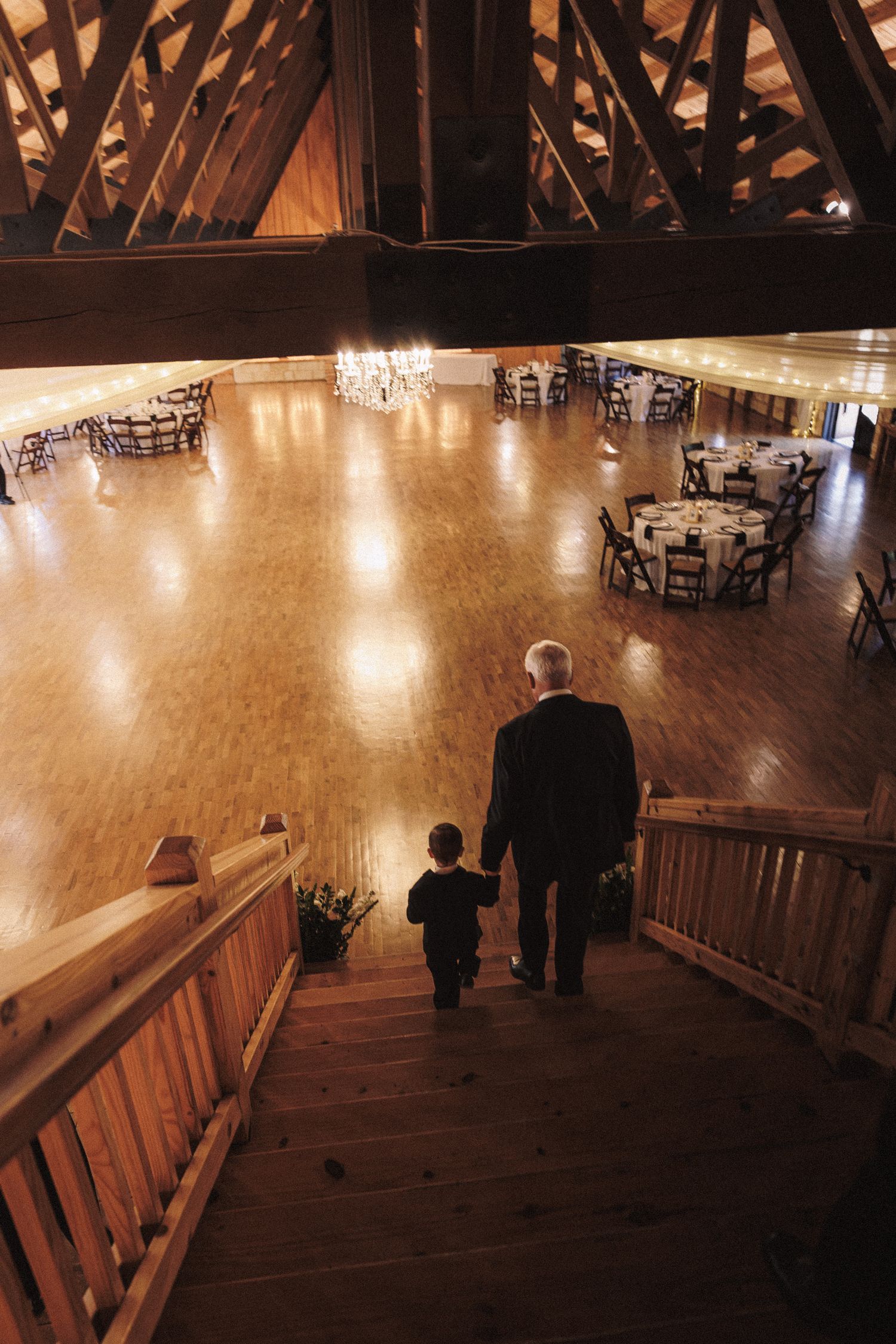 Aerial view of man and child walking into warmly lit rustic barn wedding reception venue with chandelier.