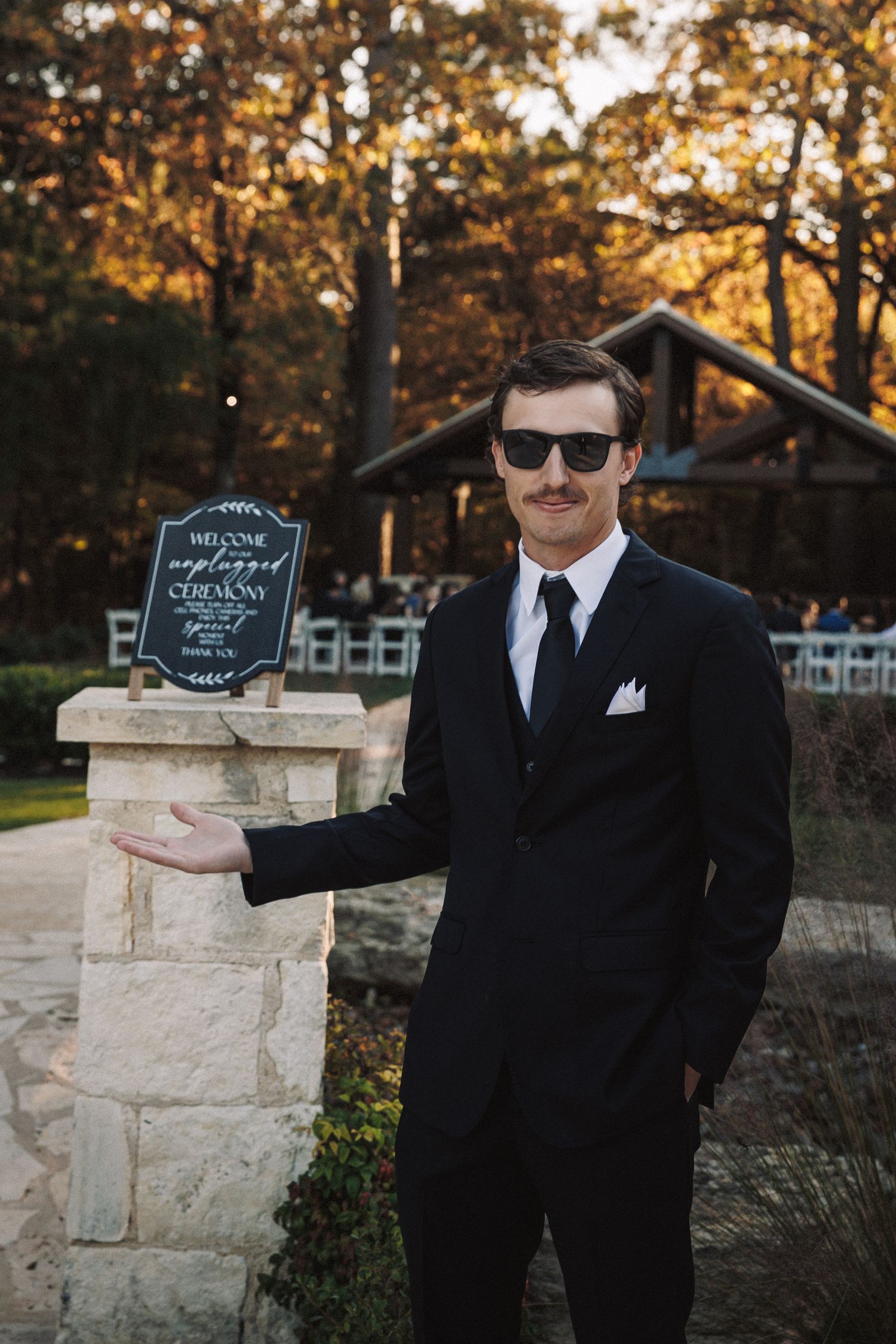 Dapper groomsman in black suit and sunglasses poses beside unplugged ceremony wedding sign at venue.