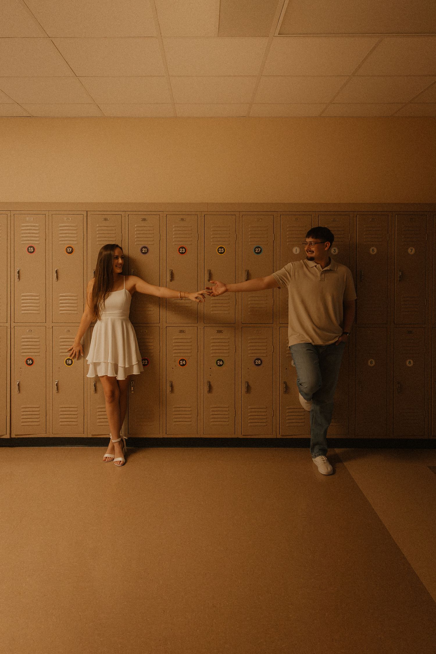 A couple holds hands at arm's length in a school hallway lined with beige lockers, she in a white dress, he in casual attire.
